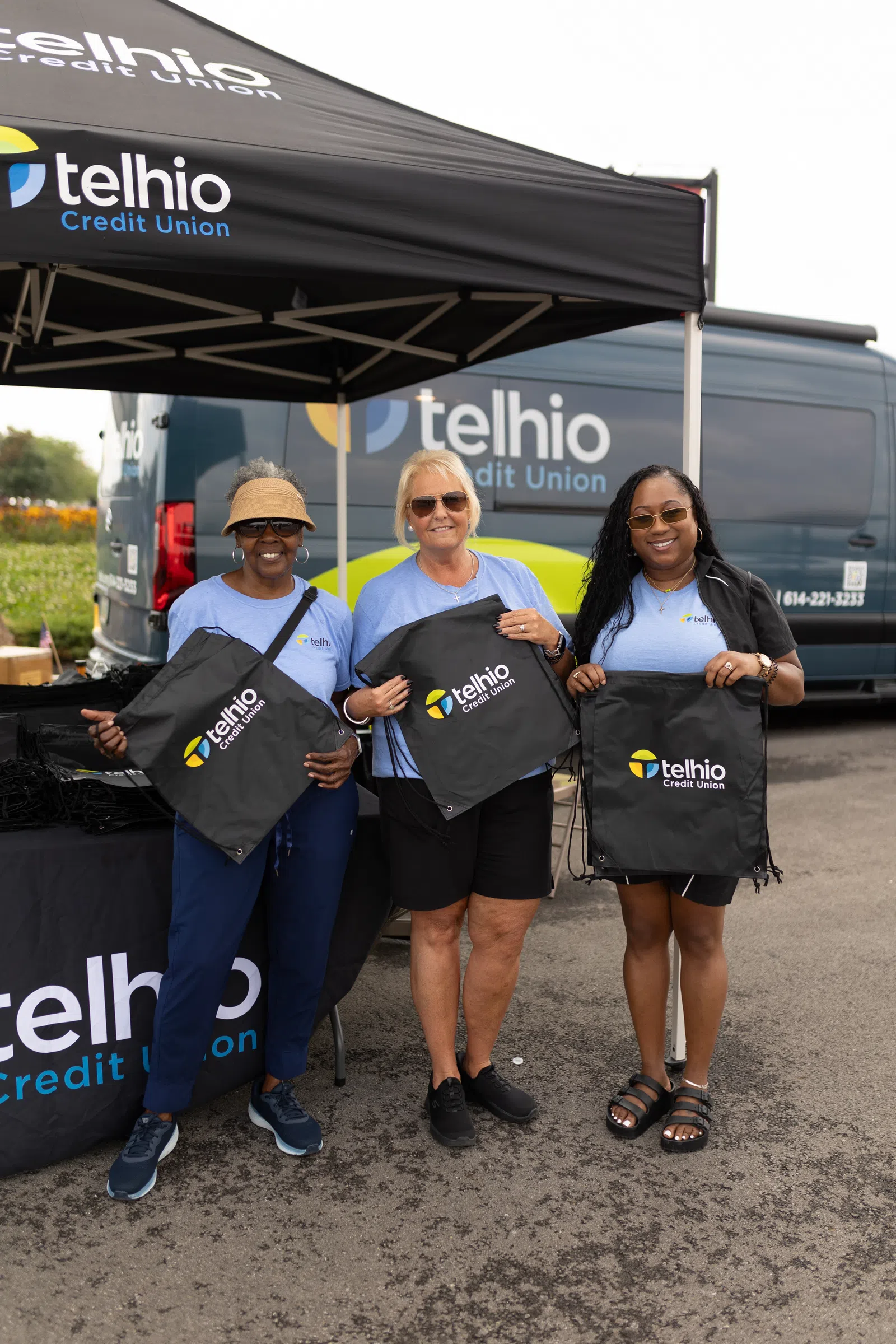 Three women in Telhio Credit Union shirts holding branded drawstring bags under a Telhio tent with a Telhio van in background