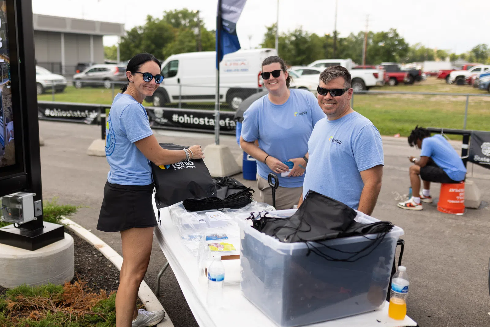 Three people in blue shirts packing black drawstring bags outdoors at an event with cars and tents in the background