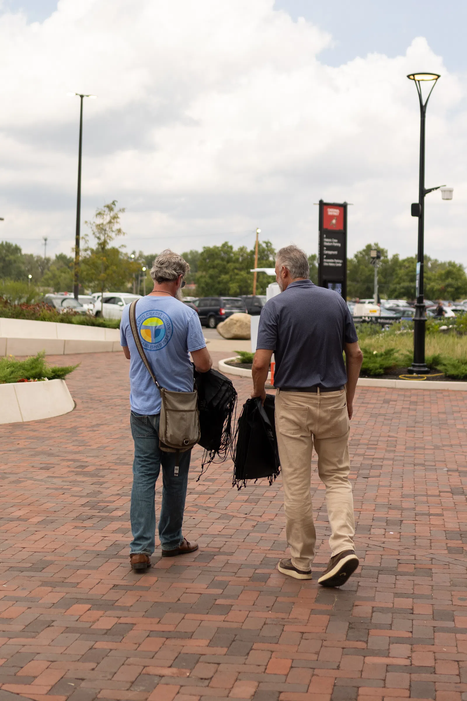 Two men walking on a brick pathway carrying folded black chairs on a cloudy day in a park-like area.