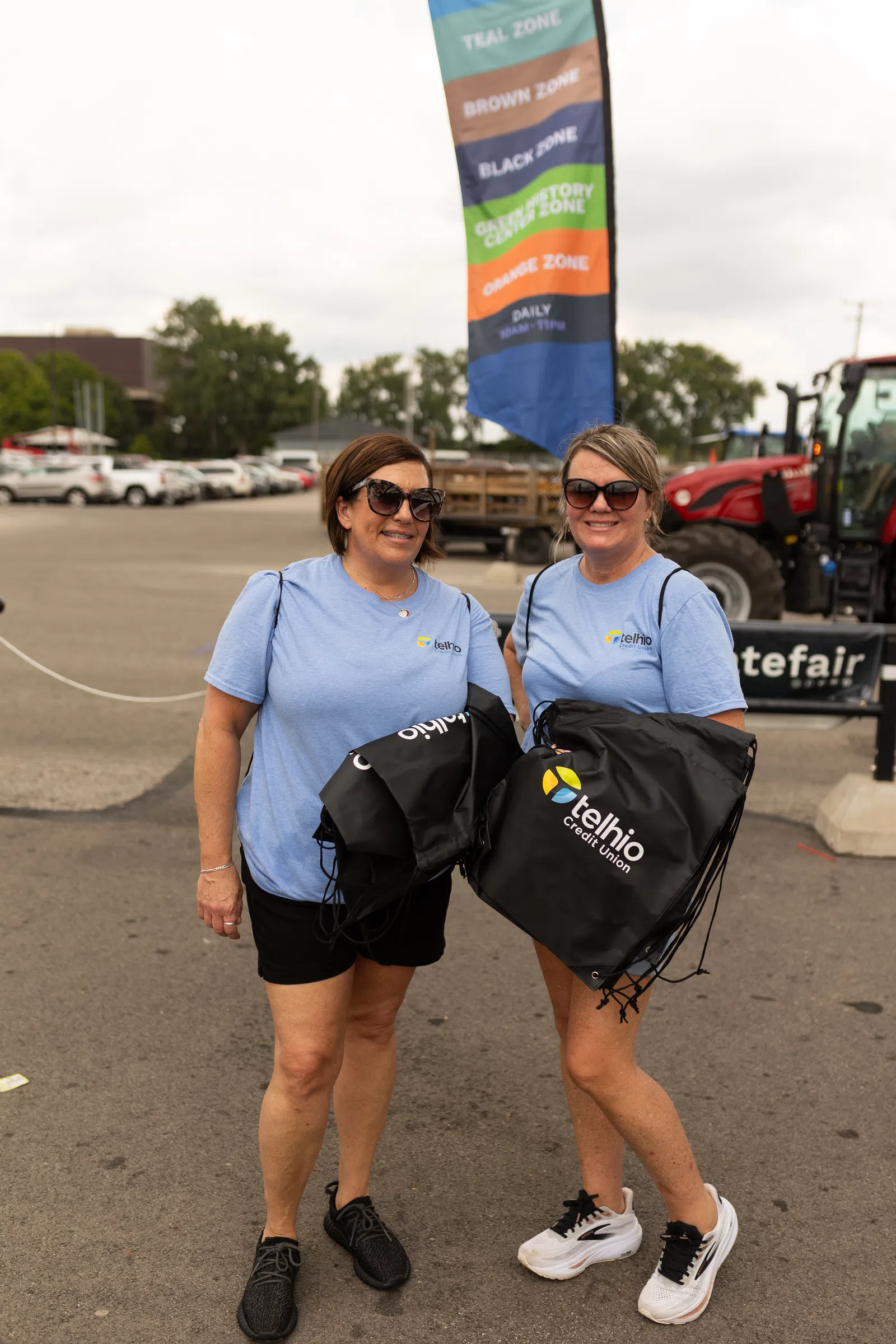 Two women wearing light blue Telhio Credit Union shirts hold branded black drawstring bags outdoors.