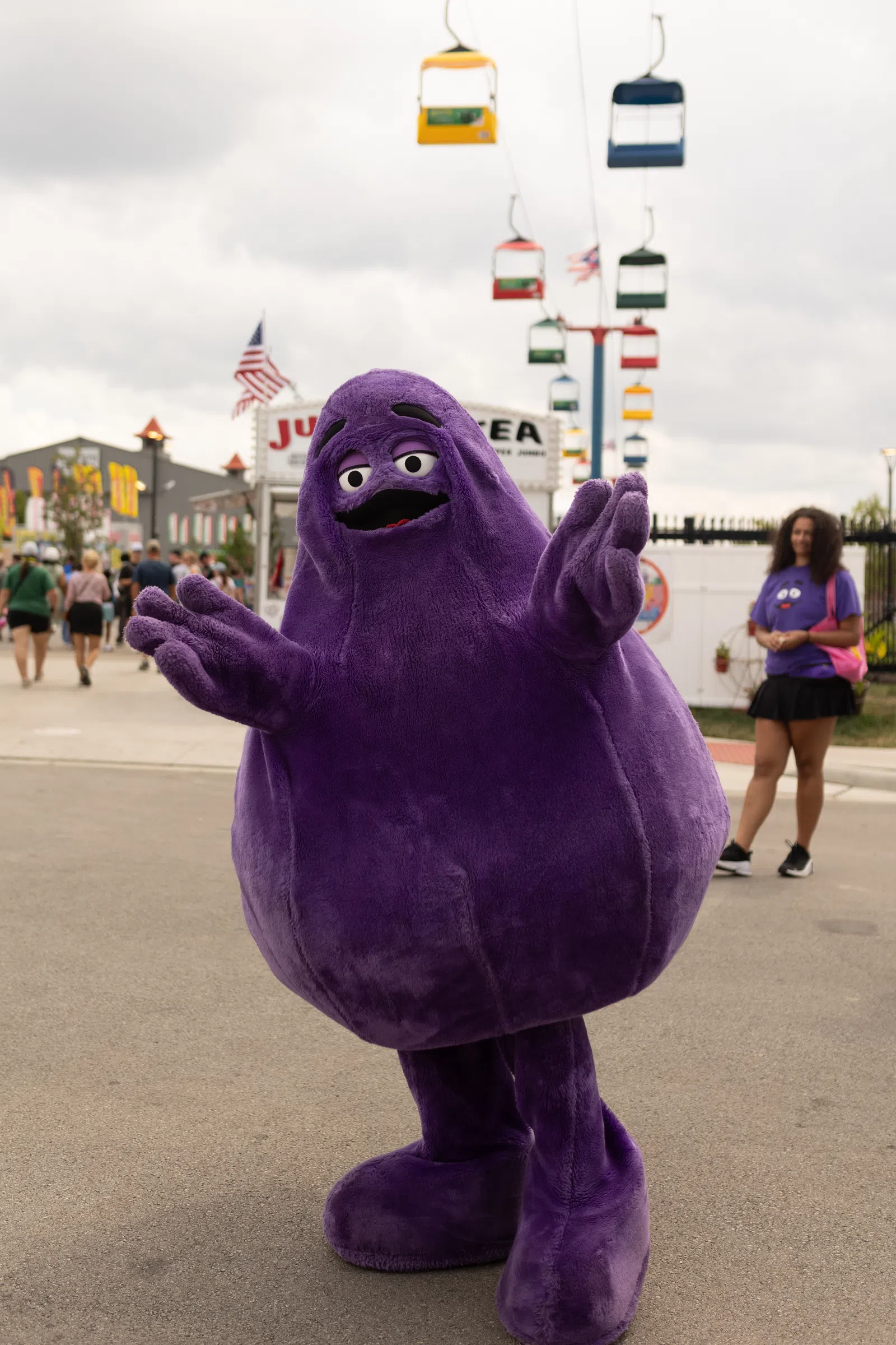 Person in a large purple mascot costume posing at an outdoor fair with colorful cable cars and people in the background