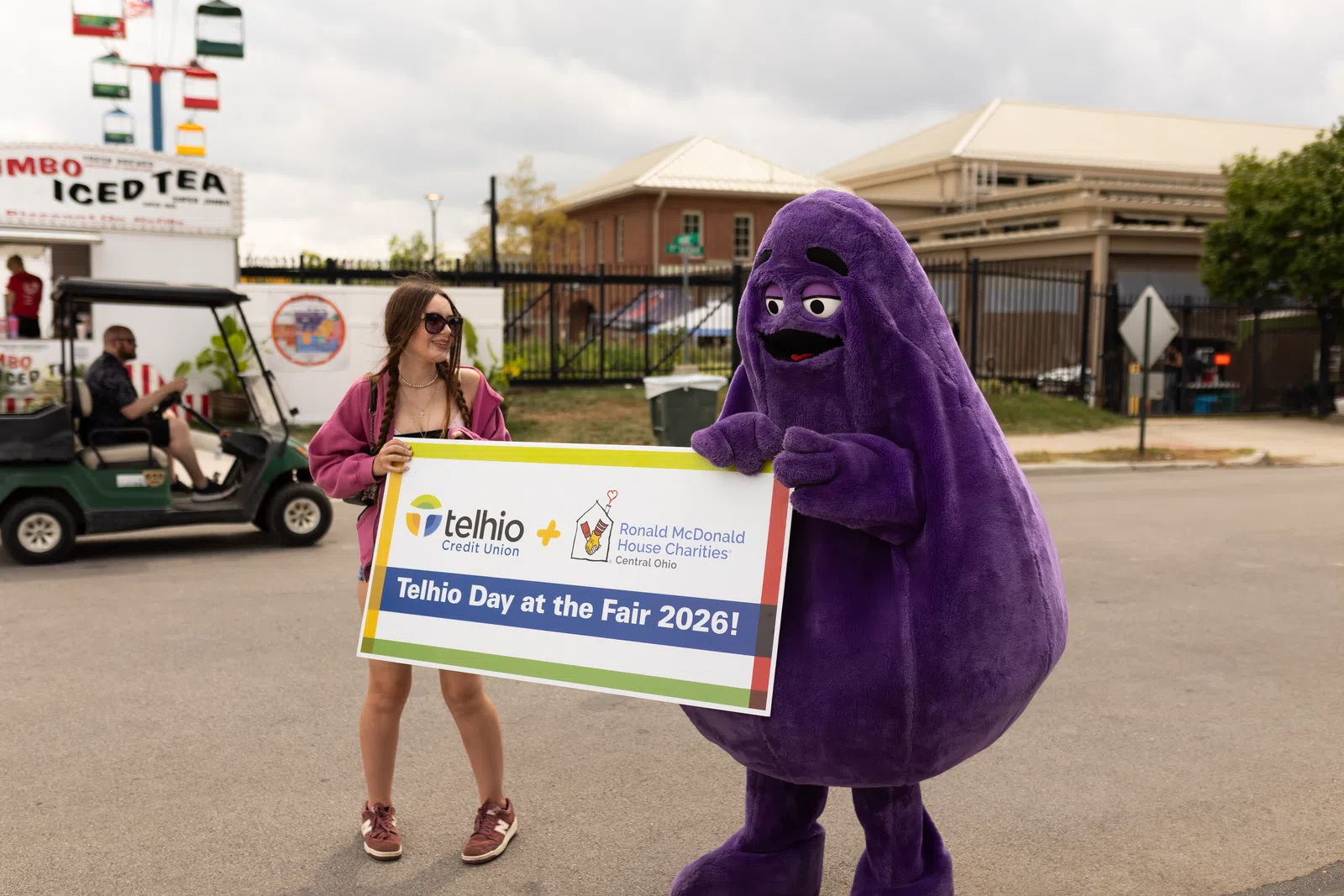 Woman with sunglasses and purple mascot holding a sign for Telhio Day at the Fair 2026 outdoors.