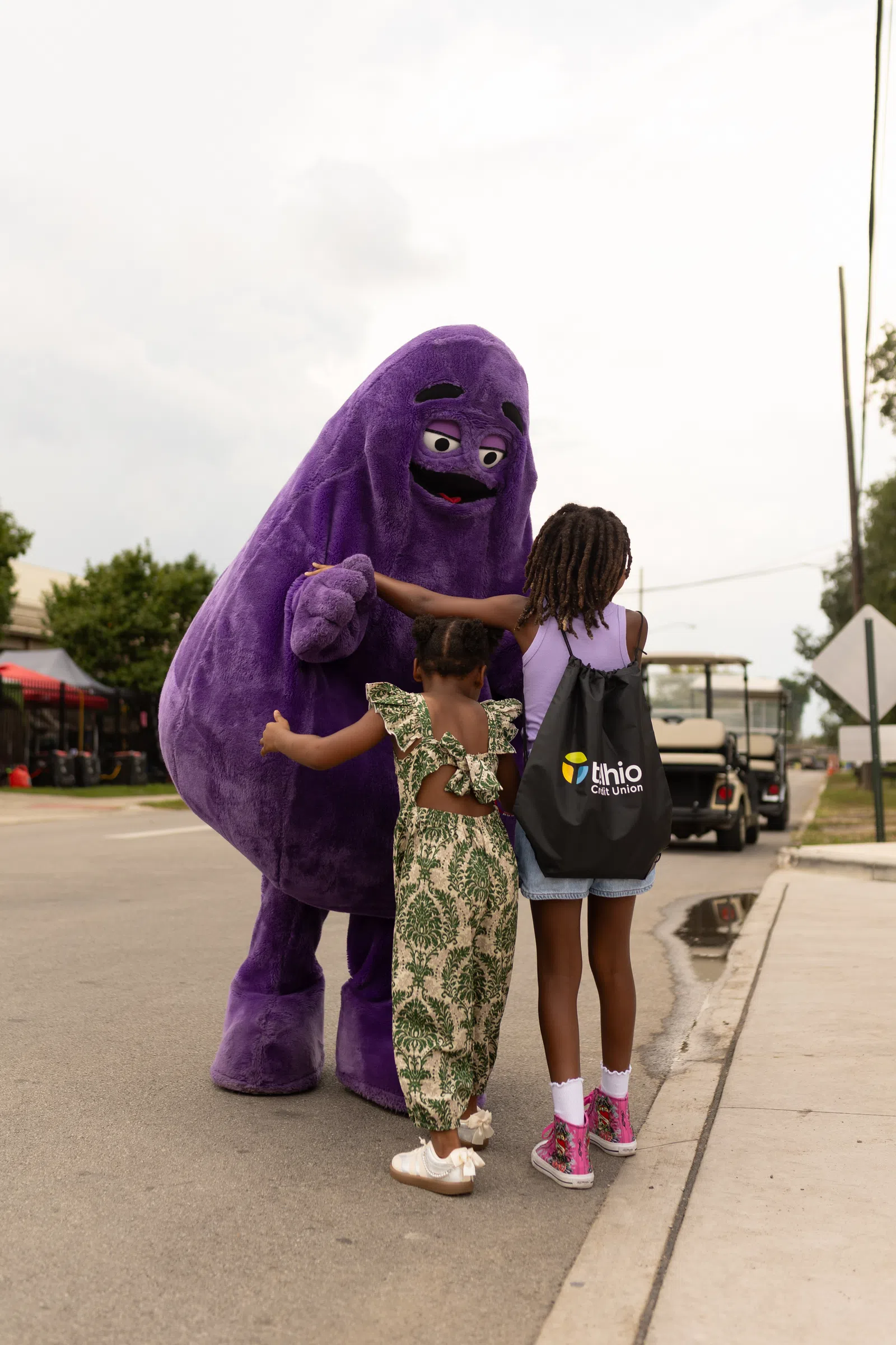 Two children interacting with a person in a large purple mascot costume on a street during daytime.