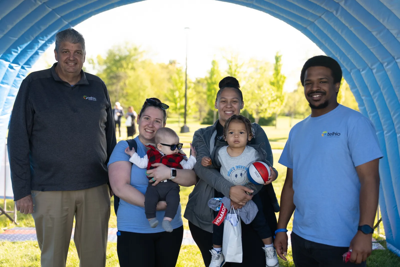 Group of smiling adults and children posing outdoors under a blue inflatable arch on a sunny day