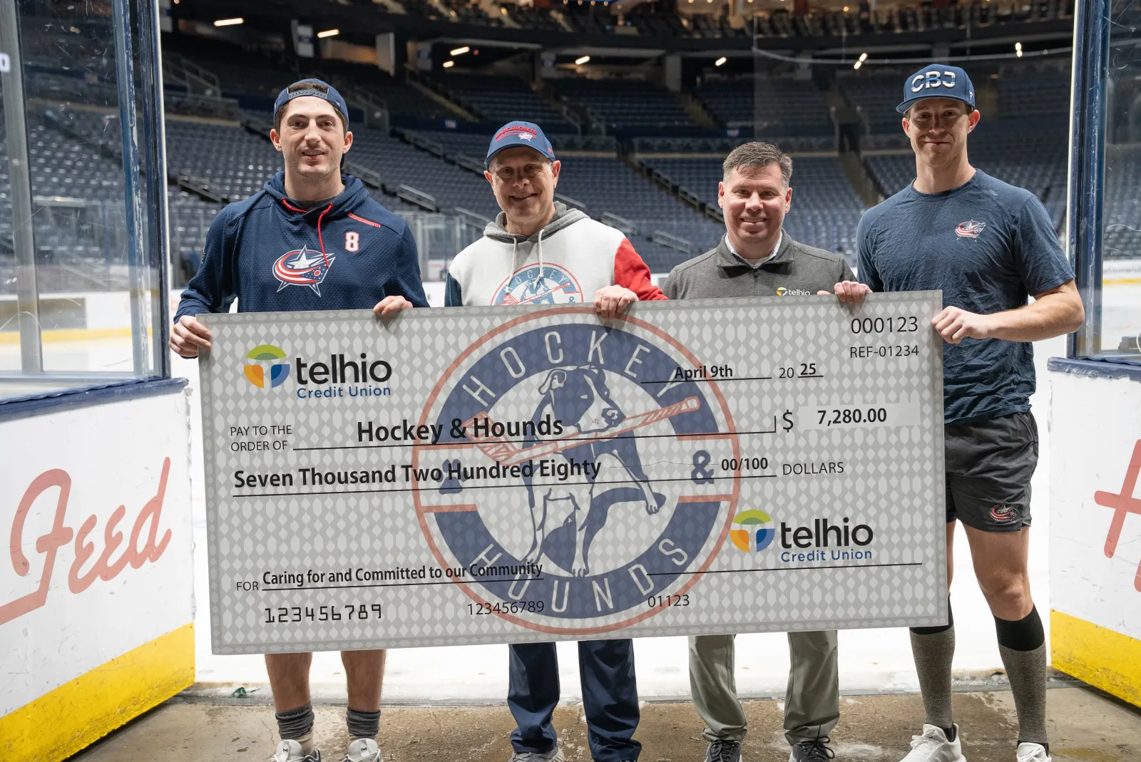 Four men on an ice hockey rink holding a large Telhio Credit Union check for $7,280 made out to Hockey & Hounds.