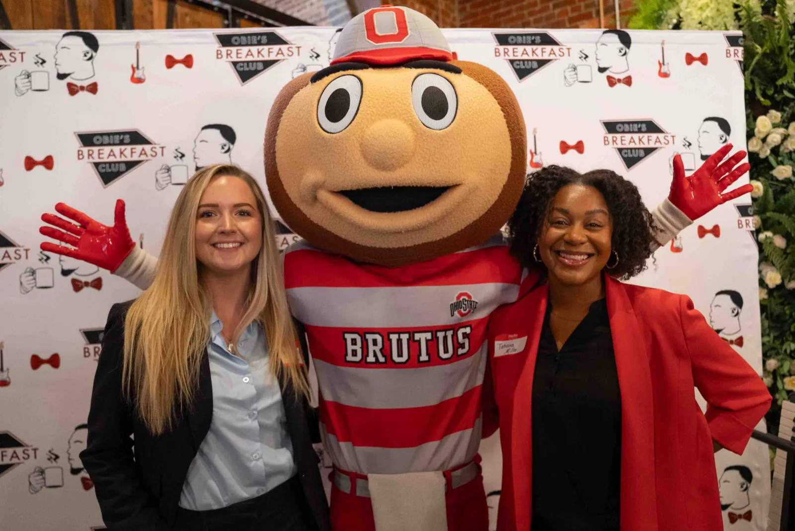 Two women smiling with Ohio State mascot Brutus Buckeye at Obie's Breakfast Club event backdrop.