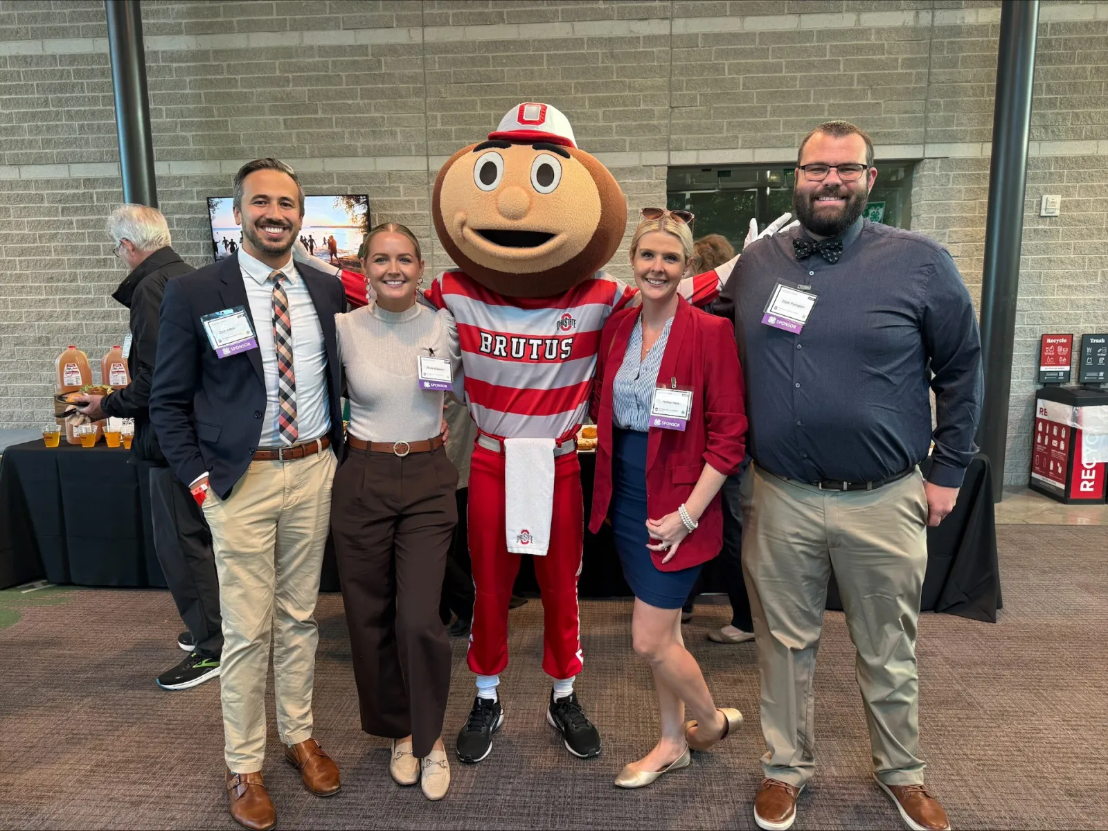 Group of four people posing with Brutus Buckeye mascot at an indoor event with brick walls and tables.