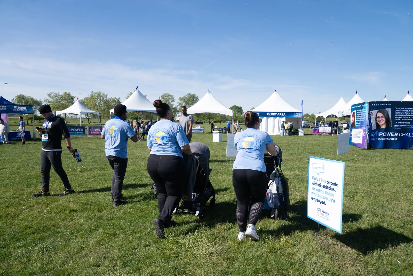 Volunteers in blue shirts push strollers on grass at an outdoor event with white tents on a sunny day.