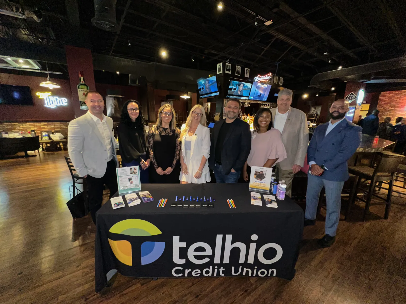 Group of professionals standing behind a Telhio Credit Union promotional table in a bar setting.