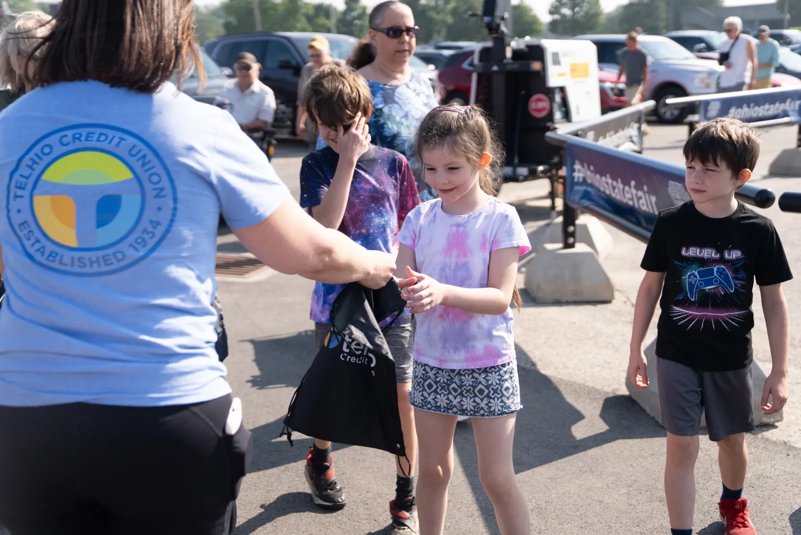 Children receiving giveaway bags from Telhio Credit Union staff during an outdoor event on a sunny day.