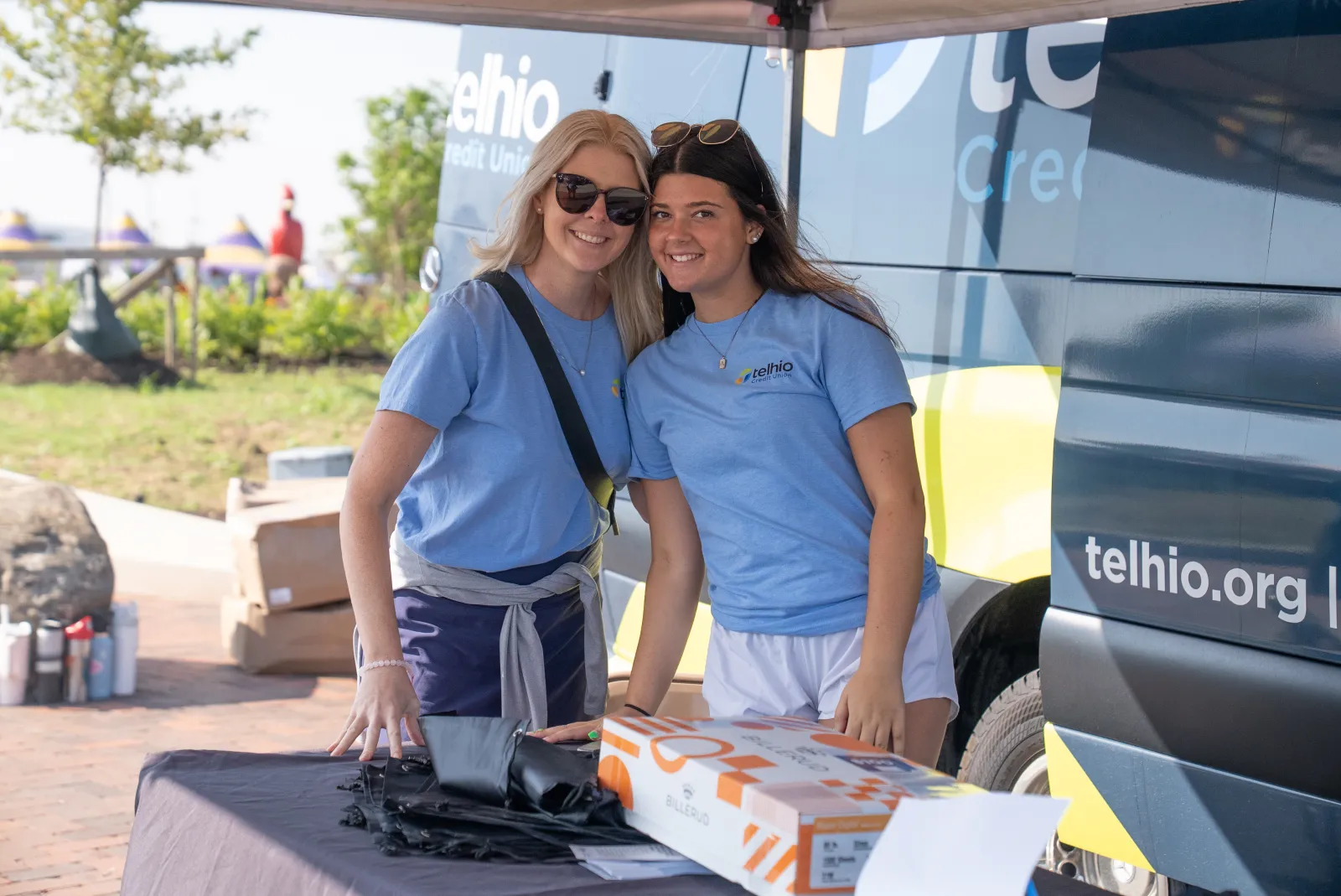 Two women in blue Telhio Credit Union shirts smiling at an outdoor event booth with branded van in background.