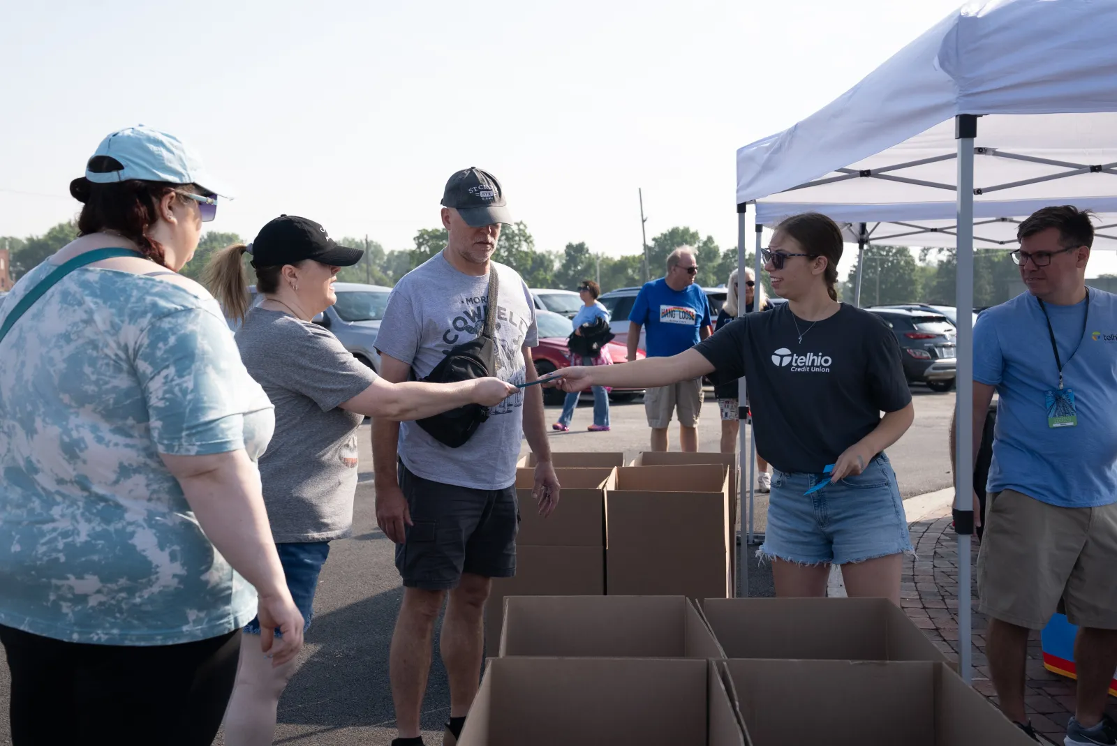 Volunteers distributing items to people at an outdoor event under a white tent with cardboard boxes