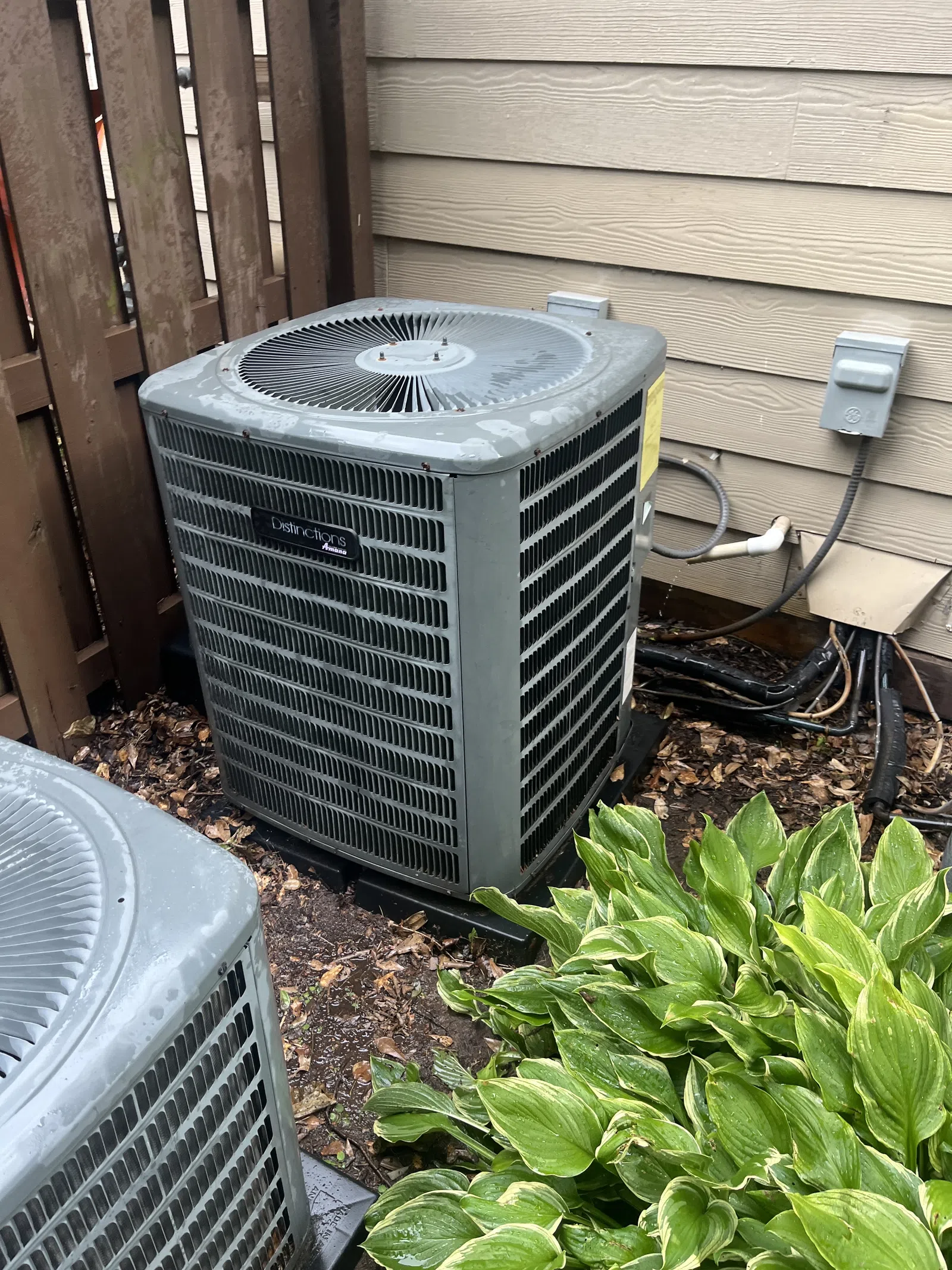 AC Maintenance, Cleaning and Tune-up in Johns Creek by The Air Company of GA.  Two outdoor air conditioning units near a wooden fence and siding with green leafy plants in front.