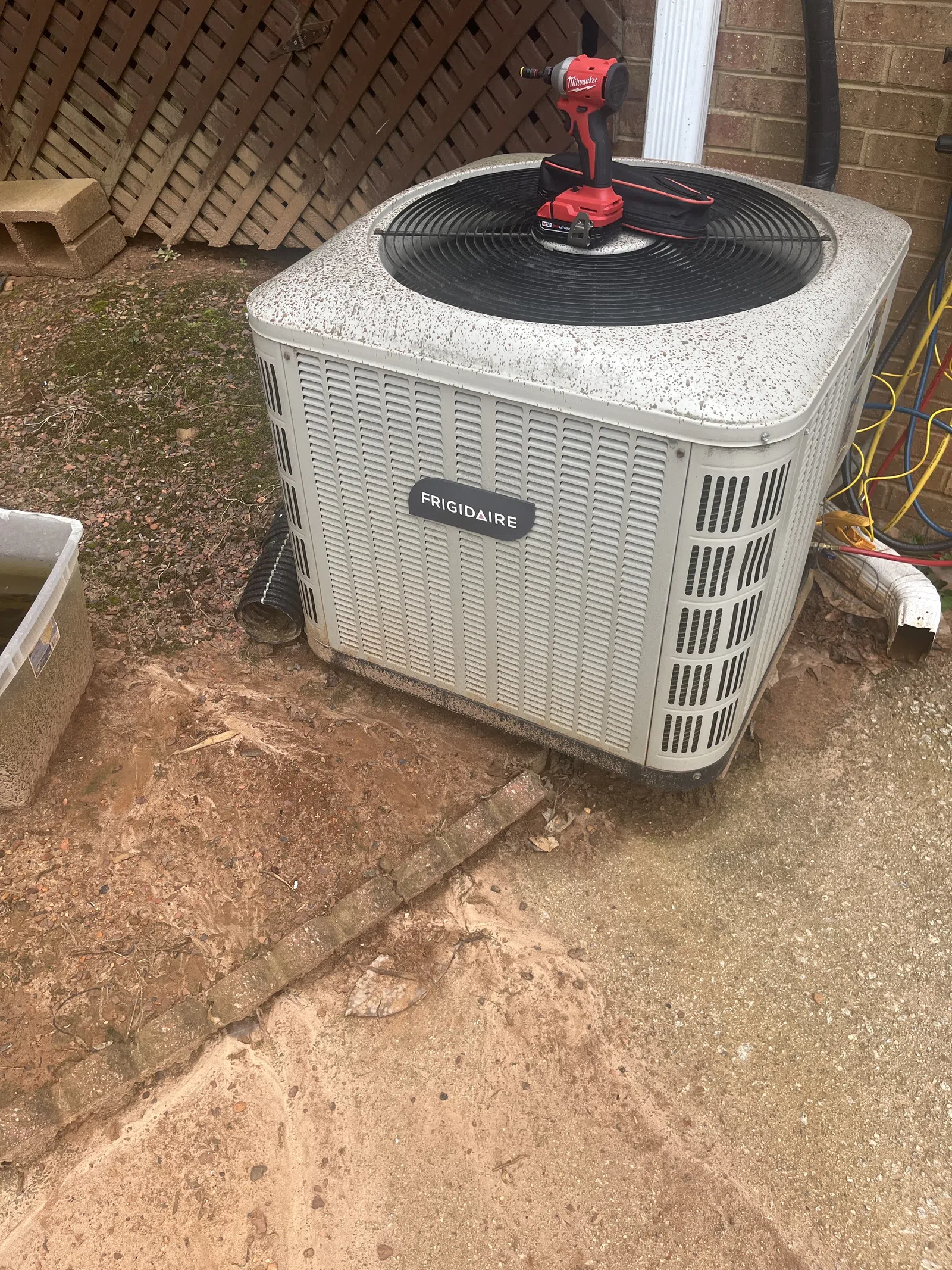 AC Preventative Maintenance in Tucker, Atlanta by The Air Company of GA. Outdoor Frigidaire air conditioner unit with a red Milwaukee drill on top beside a brick wall and garden area.
