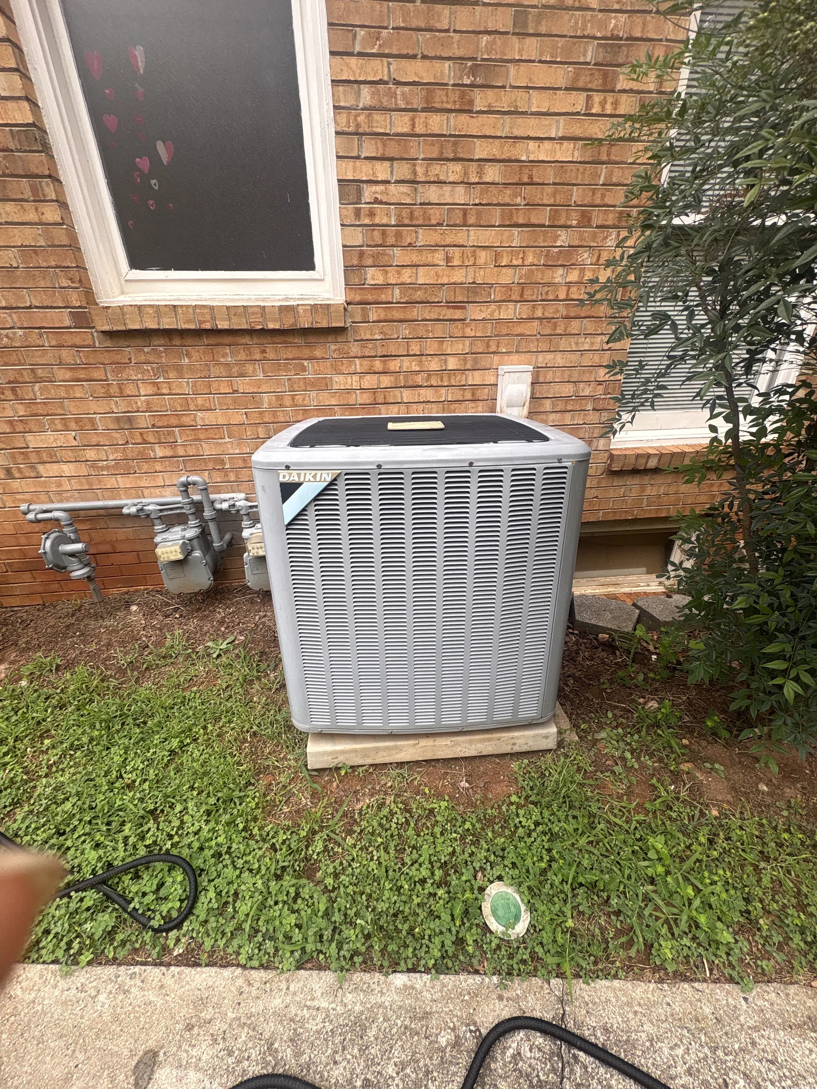 AC Repairs in Tucker Atlanta by The Air Company of GA. Outdoor Daikin air conditioning unit installed beside brick house wall with grass and shrubbery.