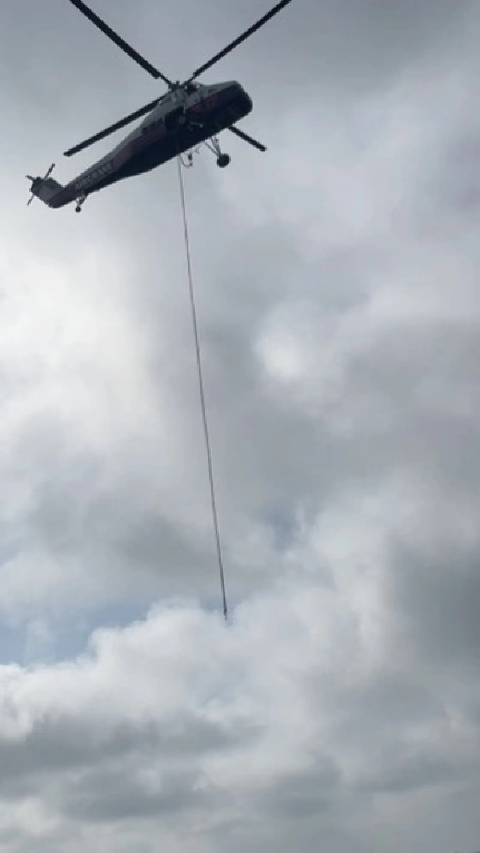 Helicopter flying in cloudy sky with a long cable hanging down vertically from it.