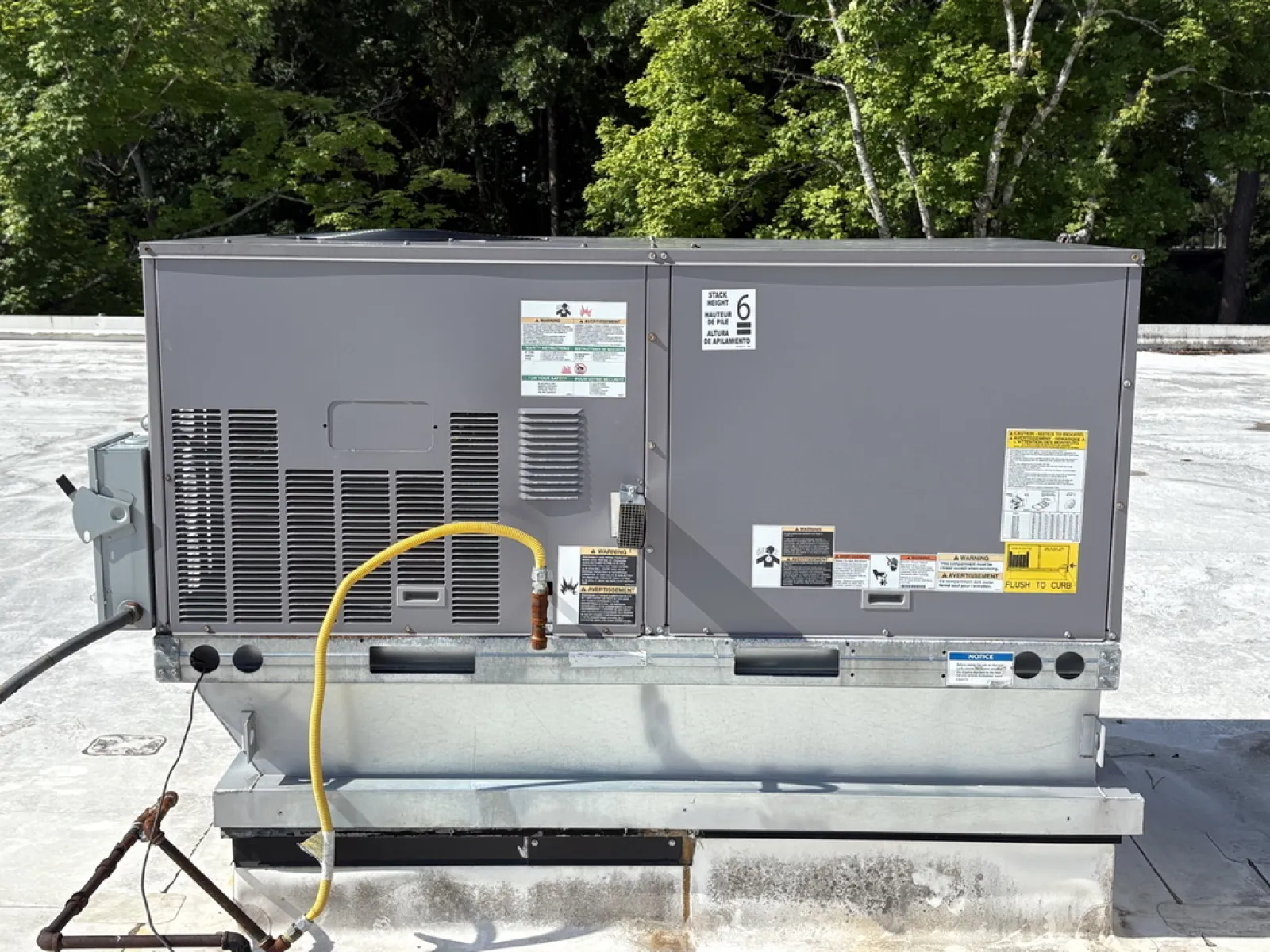 Commercial rooftop HVAC unit with gray metal casing and yellow gas line on flat roof with trees in background