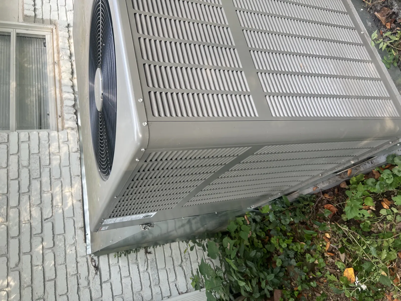 Outdoor air conditioning unit installed next to a brick house wall with surrounding greenery
