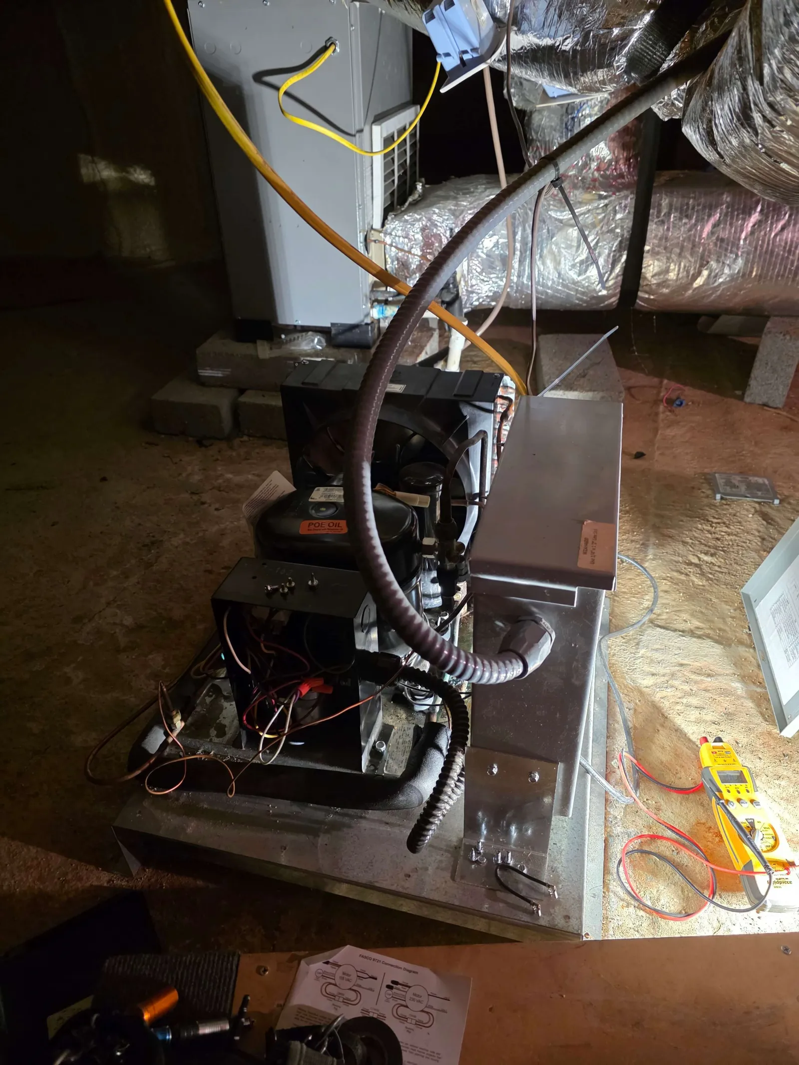 Open HVAC system unit with exposed wiring and ductwork in basement under repair with testing tool nearby.