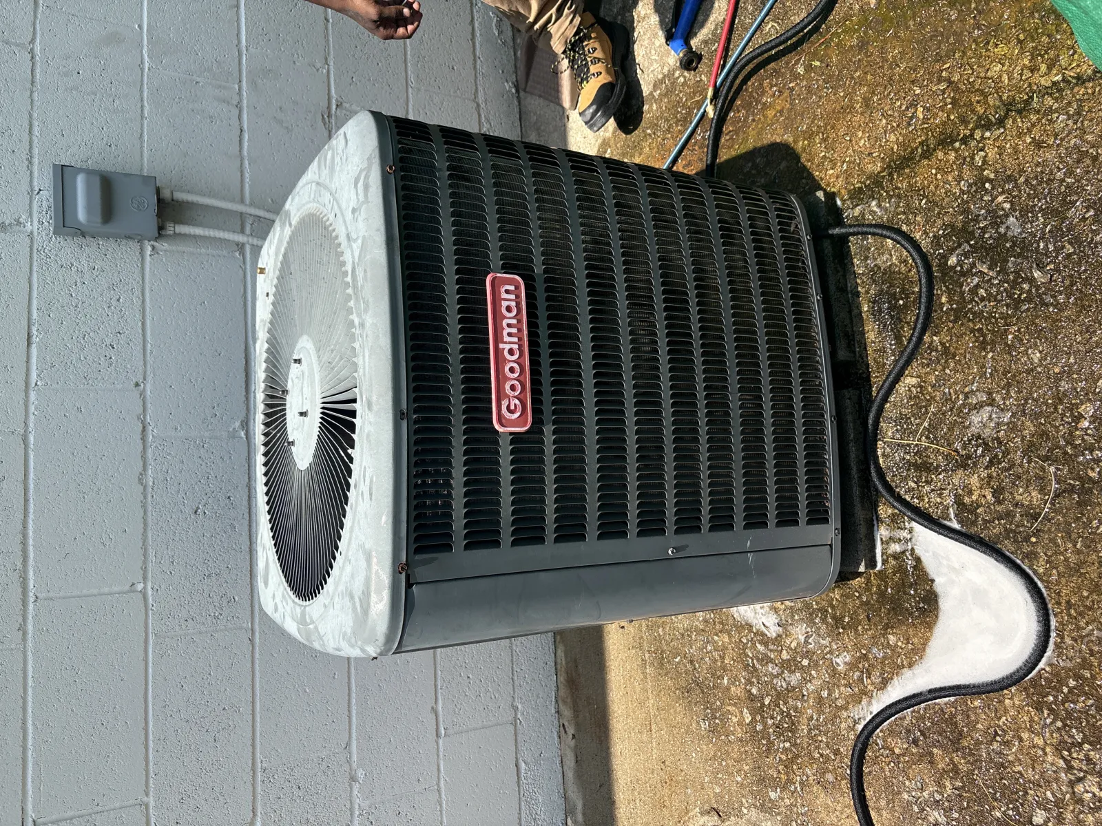 Goodman air conditioning unit installed outside a building with visible power cable and technician's boots nearby
