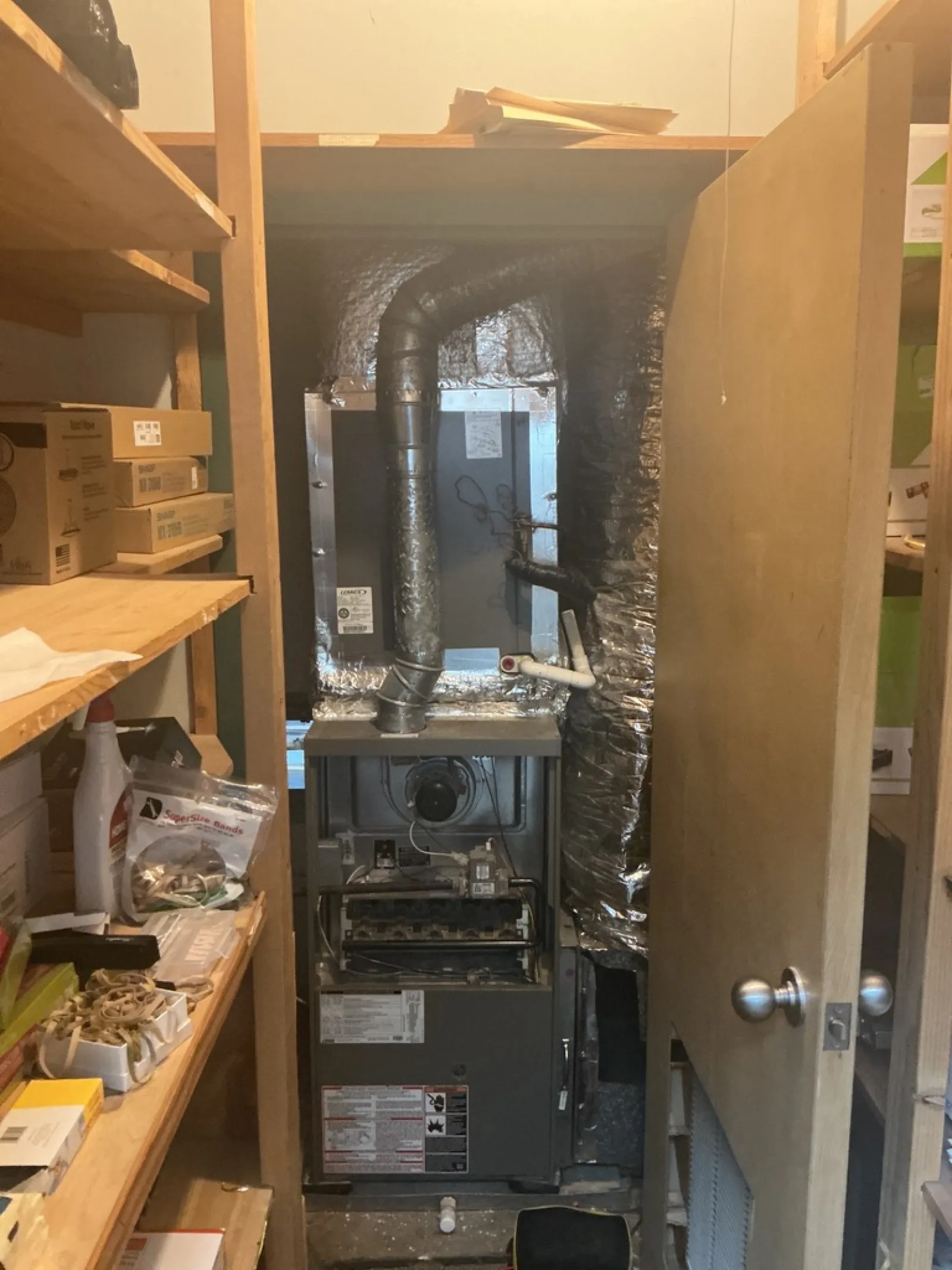 Heating furnace unit installed in a small utility closet with wooden shelves and stored items.