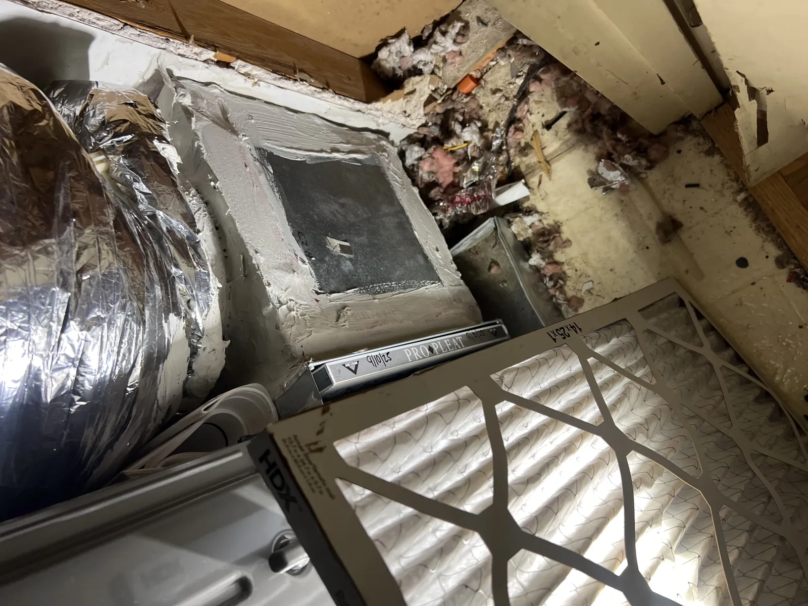 HVAC ductwork and dirty air filter in a basement with exposed insulation and dust debris. Heating system maintenance scene.