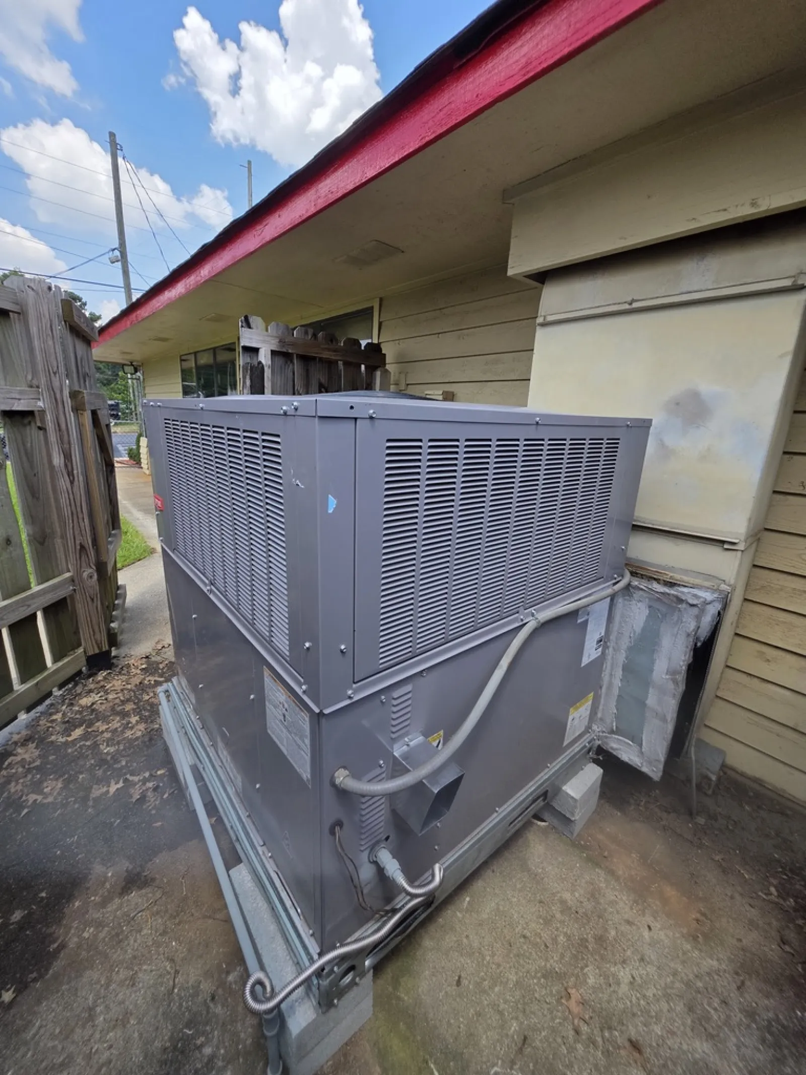 Large outdoor HVAC unit installed beside a beige building under a red-trimmed roof on concrete pad