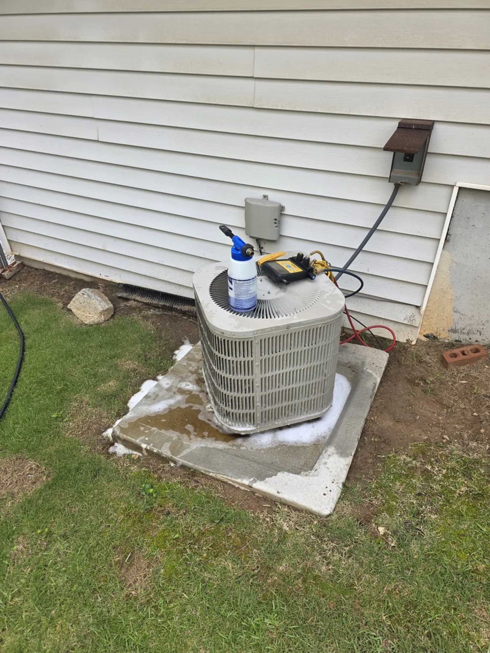 Outdoor air conditioning unit on concrete pad beside house with cleaning supplies on top and green grass surrounding.