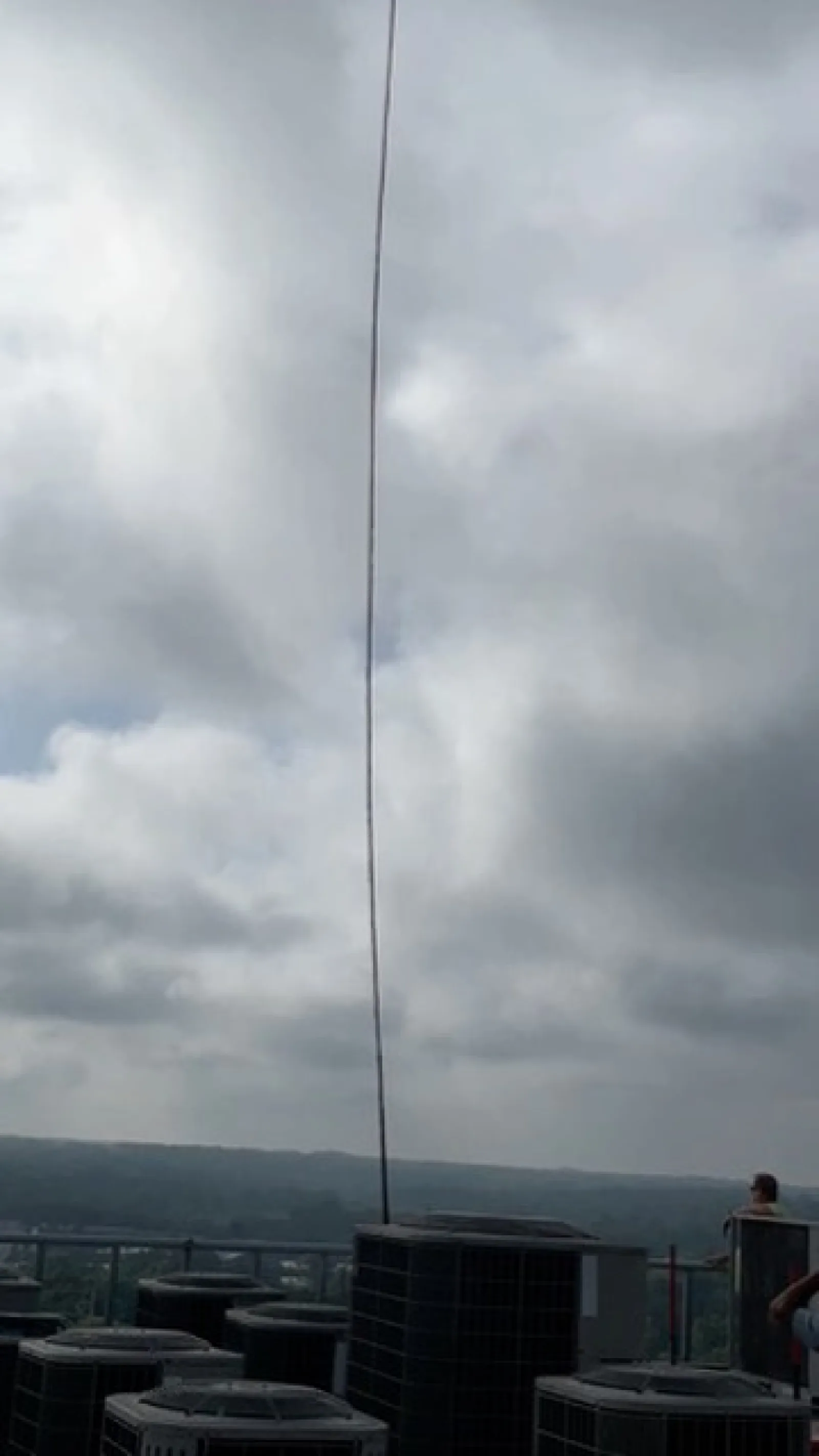 Long thin antenna mounted on rooftop with multiple HVAC units under cloudy sky in daytime