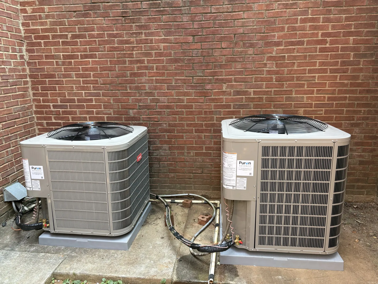 Two outdoor HVAC units installed on concrete pads against a brick wall with connected piping and wiring.