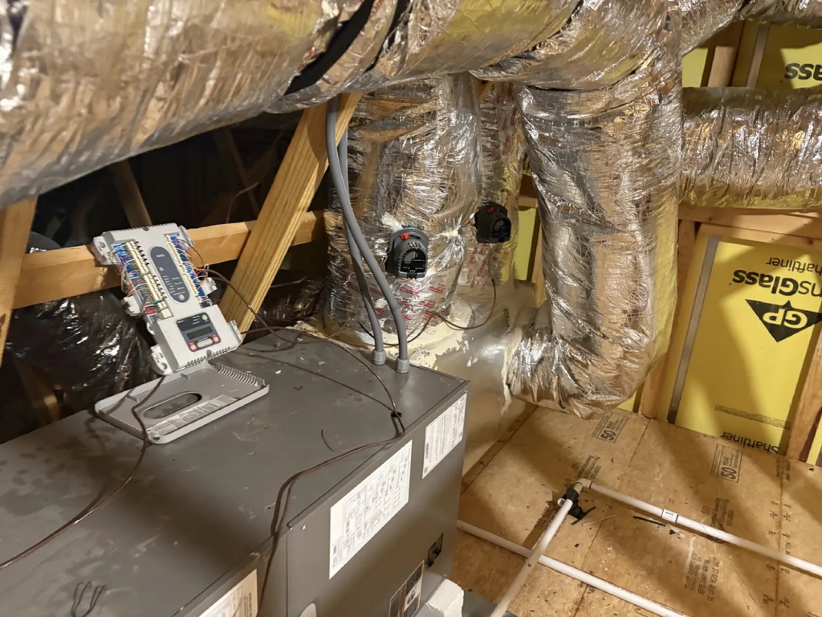 Attic HVAC system with insulated ductwork, control panel, and wooden framing in a residential home.
