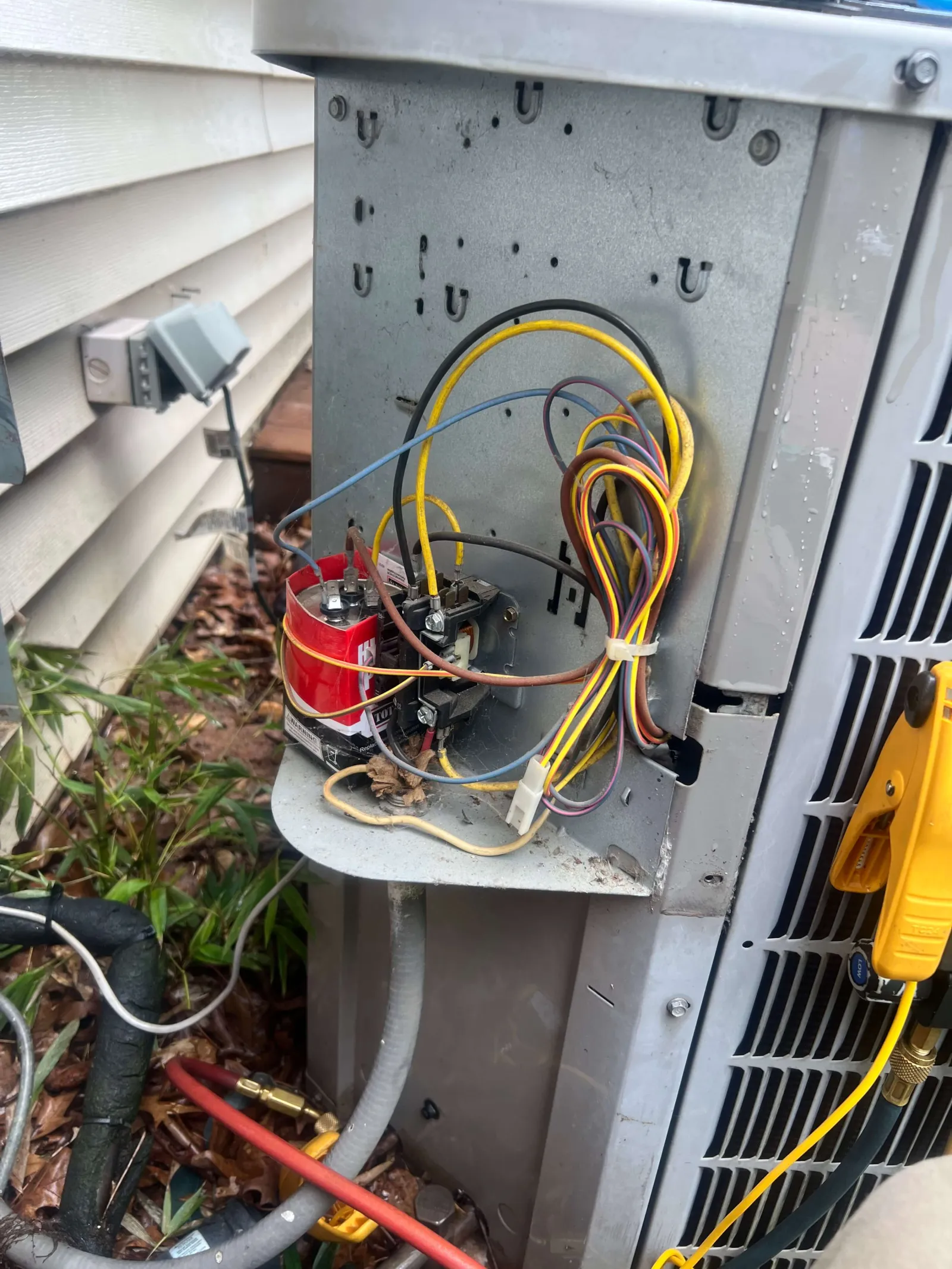 Open outdoor HVAC unit showing wires, a red capacitor, and a yellow diagnostic tool attached during maintenance.