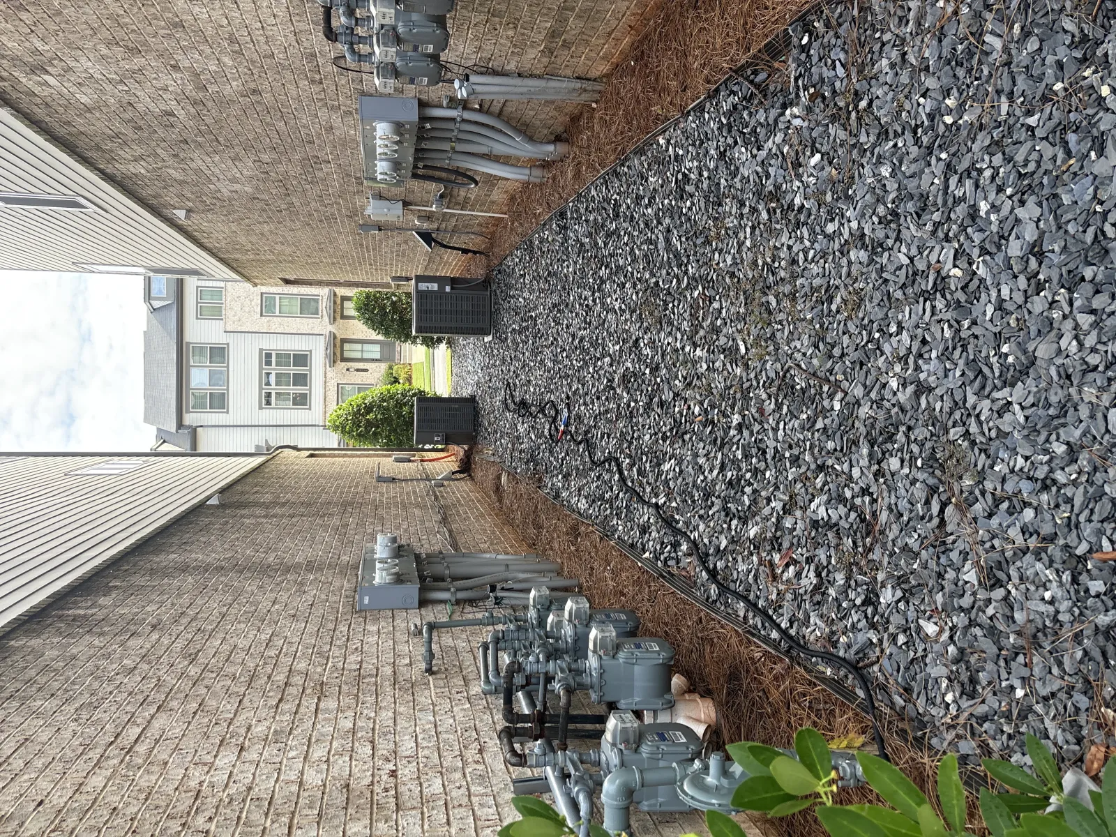 Narrow side yard between two houses with gravel ground, utility meters, and electrical equipment along the brick wall.