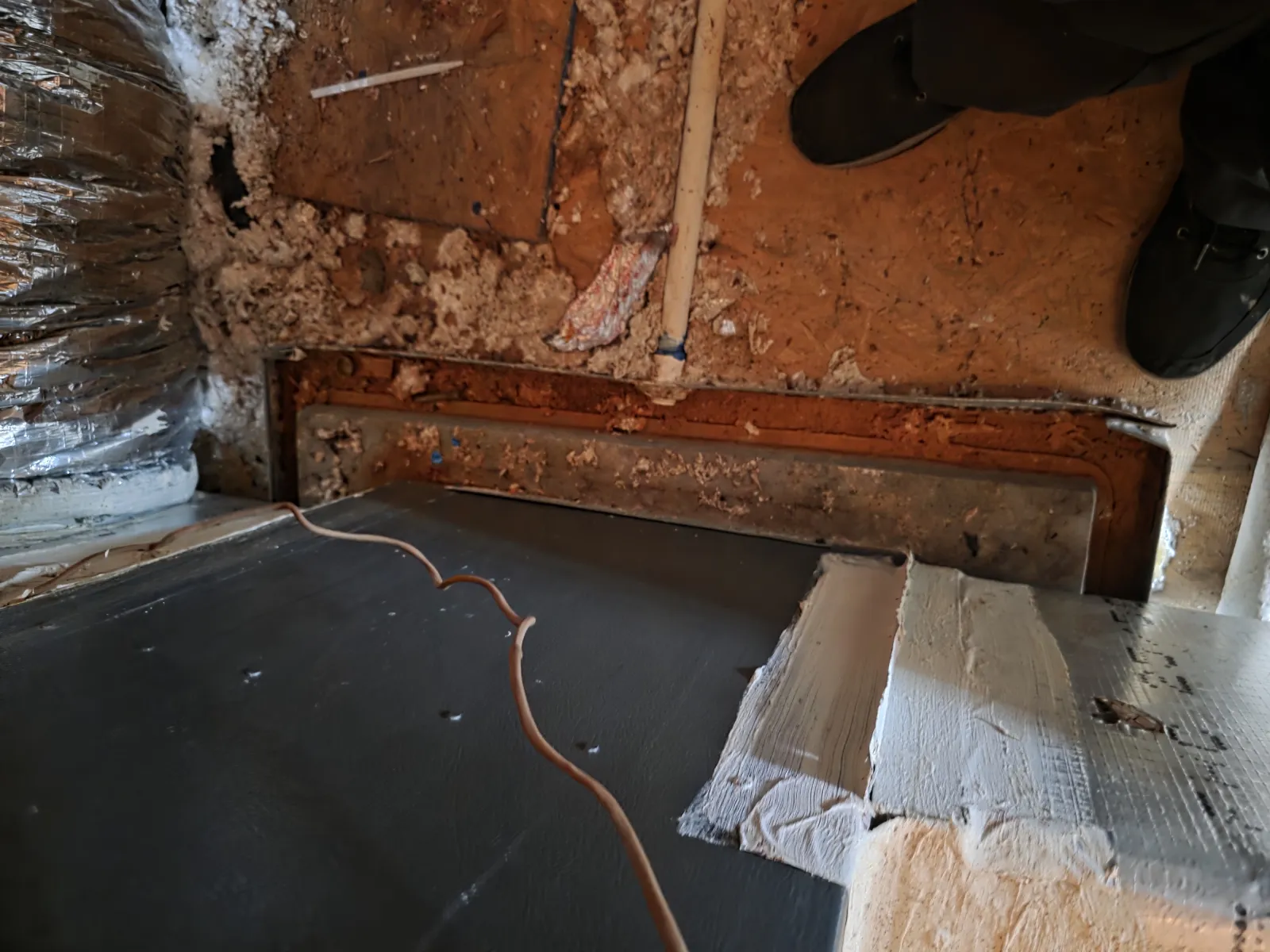 Exposed crawl space with insulation, piping, and a dark floor panel beneath a wooden subfloor with shoes visible.
