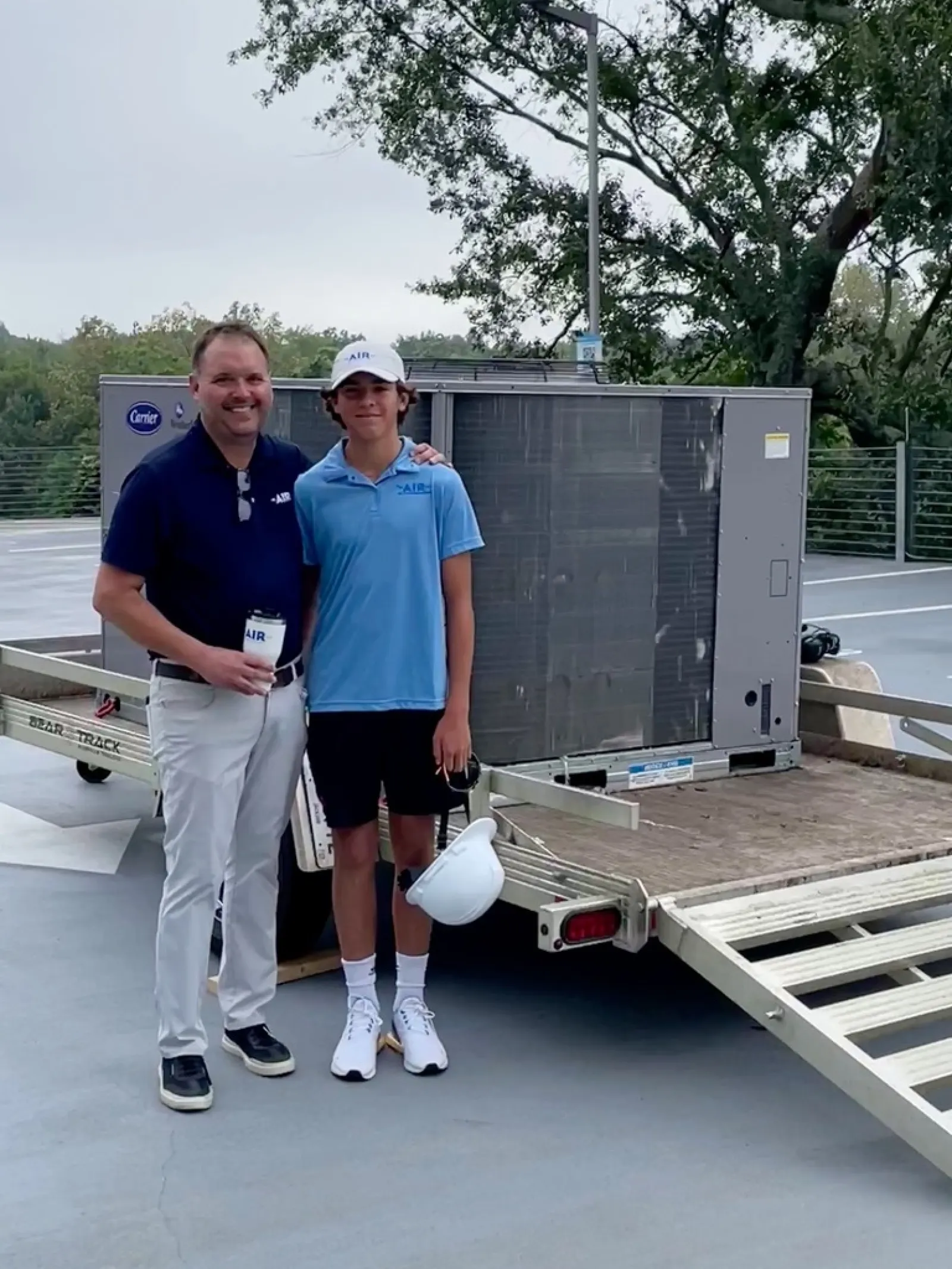 Two men in matching AIR uniforms stand next to an HVAC unit loaded on a trailer in an outdoor parking area.
