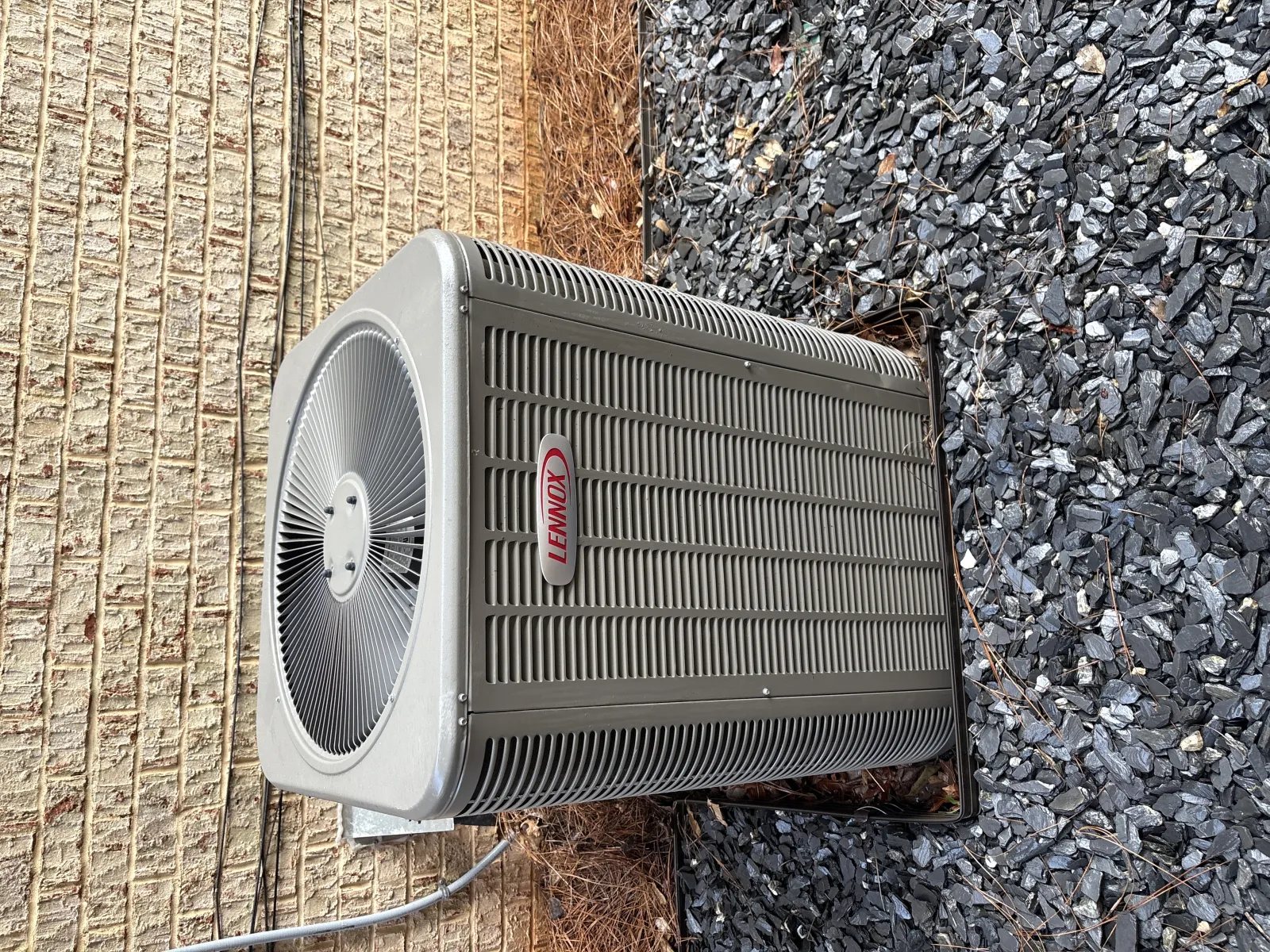 Outdoor Lennox air conditioning unit installed beside brick wall and gravel ground.