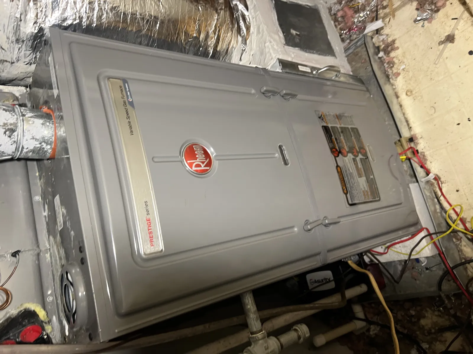Gray Rheem Prestige Series gas furnace installed in a basement with insulating foil and visible pipes.
