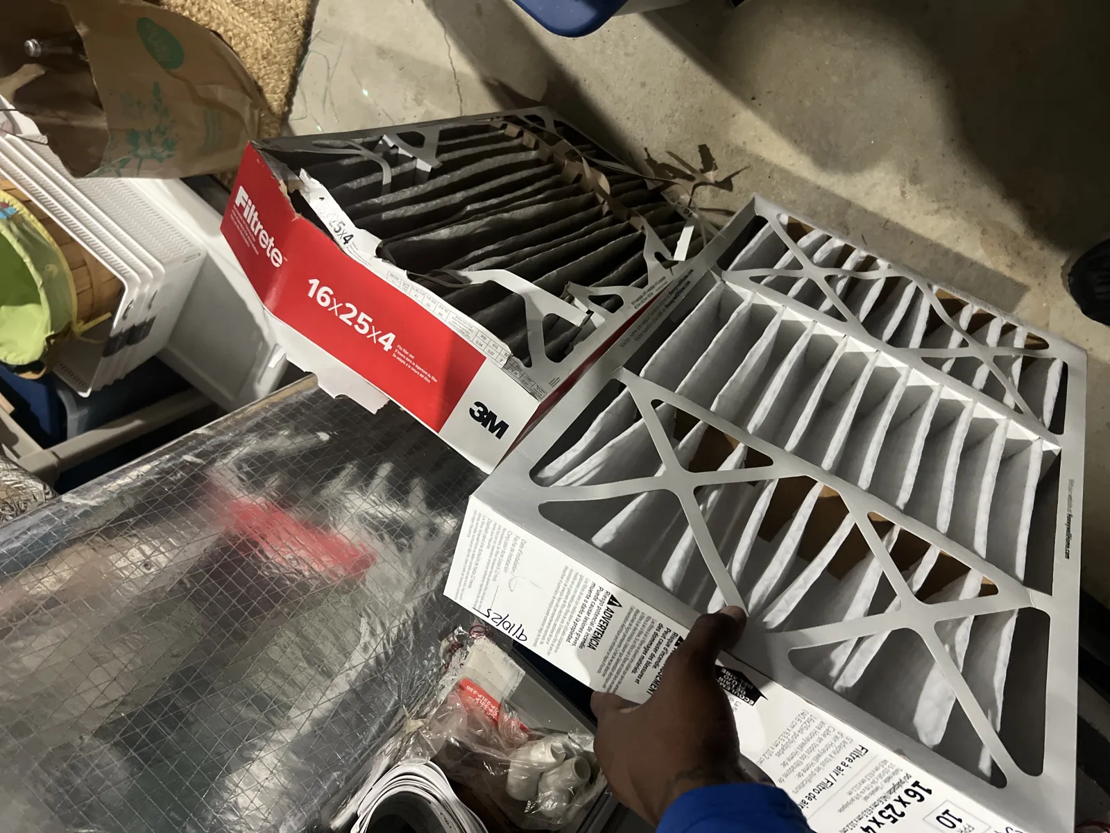 Person holding a new white 16x25x4 air filter next to a used dirty filter in a basement or storage area.