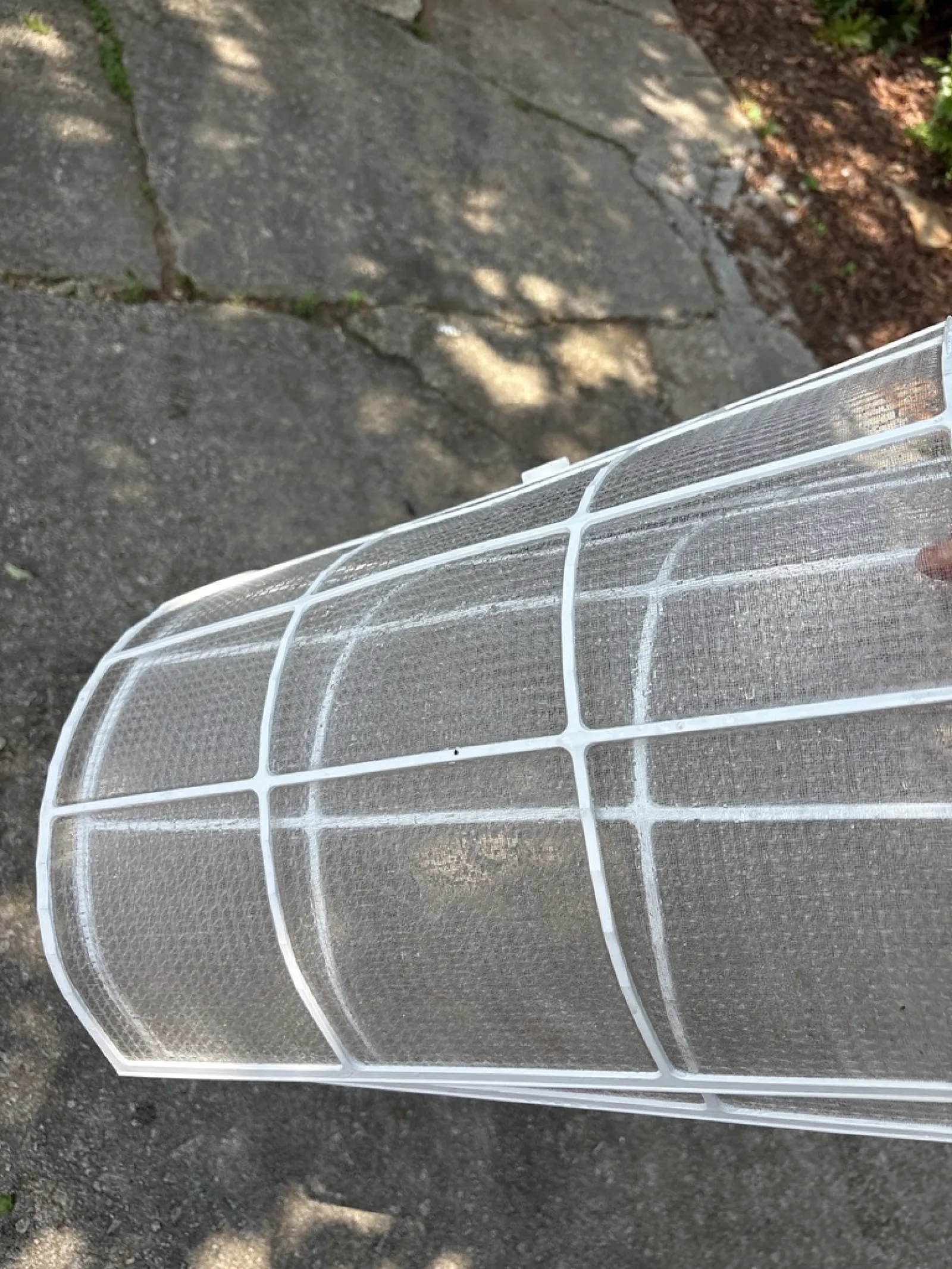 Close-up of a white plastic air conditioner filter with mesh grid held outdoors on a concrete surface.