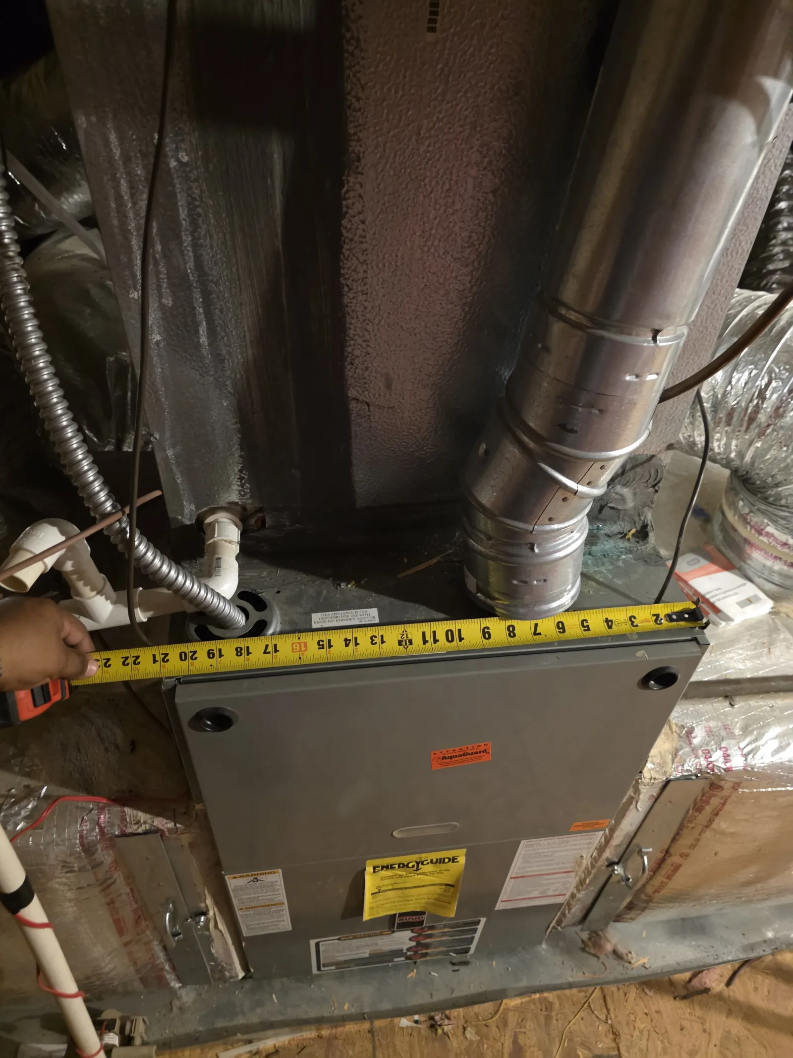 Measuring the width of a furnace unit with a tape measure showing 22 inches in a basement duct setup.
