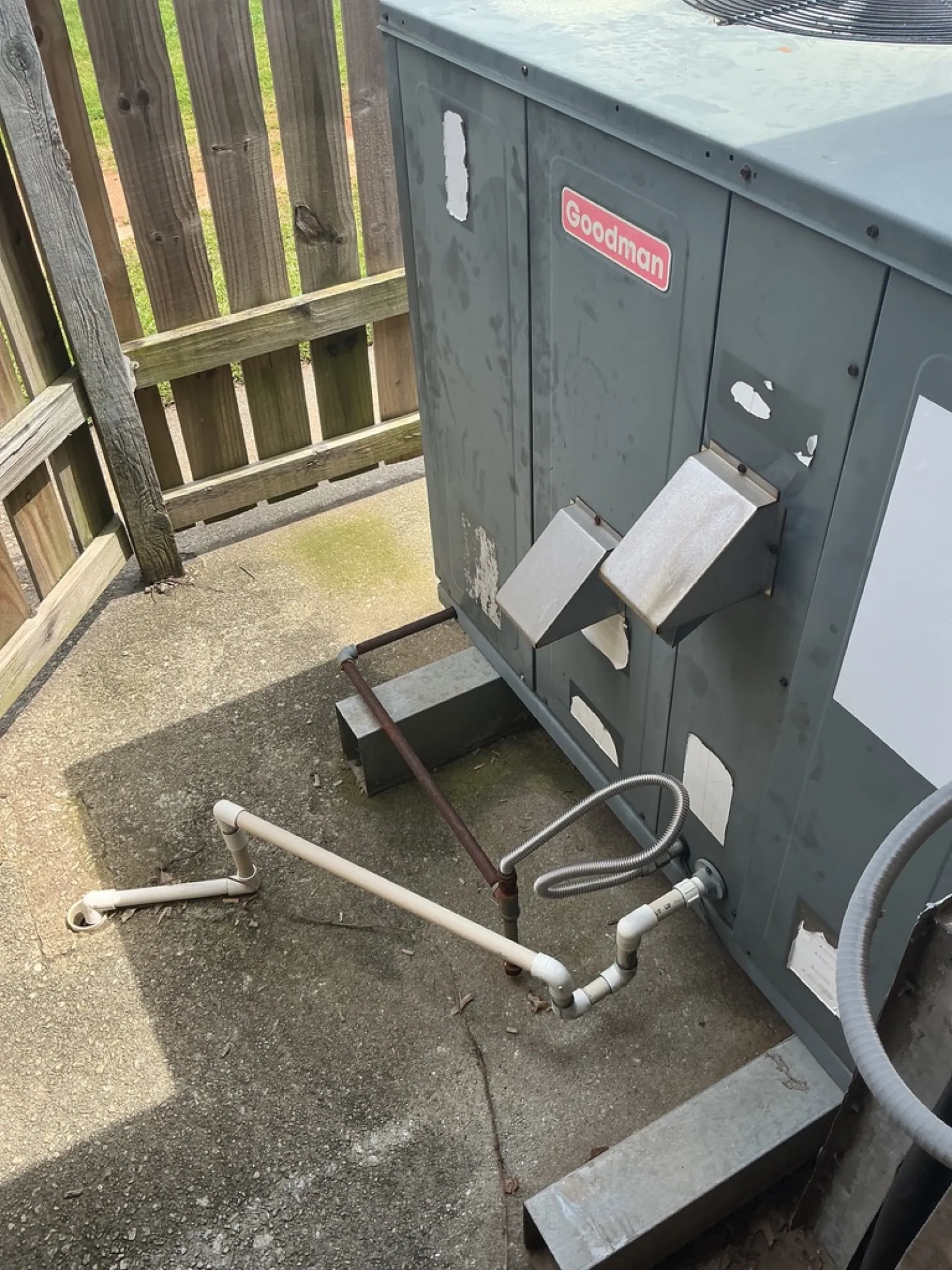Goodman HVAC unit installed outdoors with connected white and metal piping beside wooden fence on concrete ground.