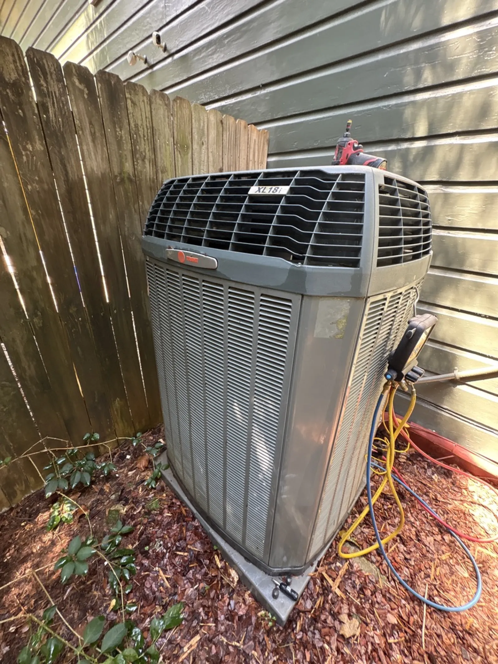 Outdoor HVAC unit installed next to house siding and wooden fence surrounded by mulch and plants.