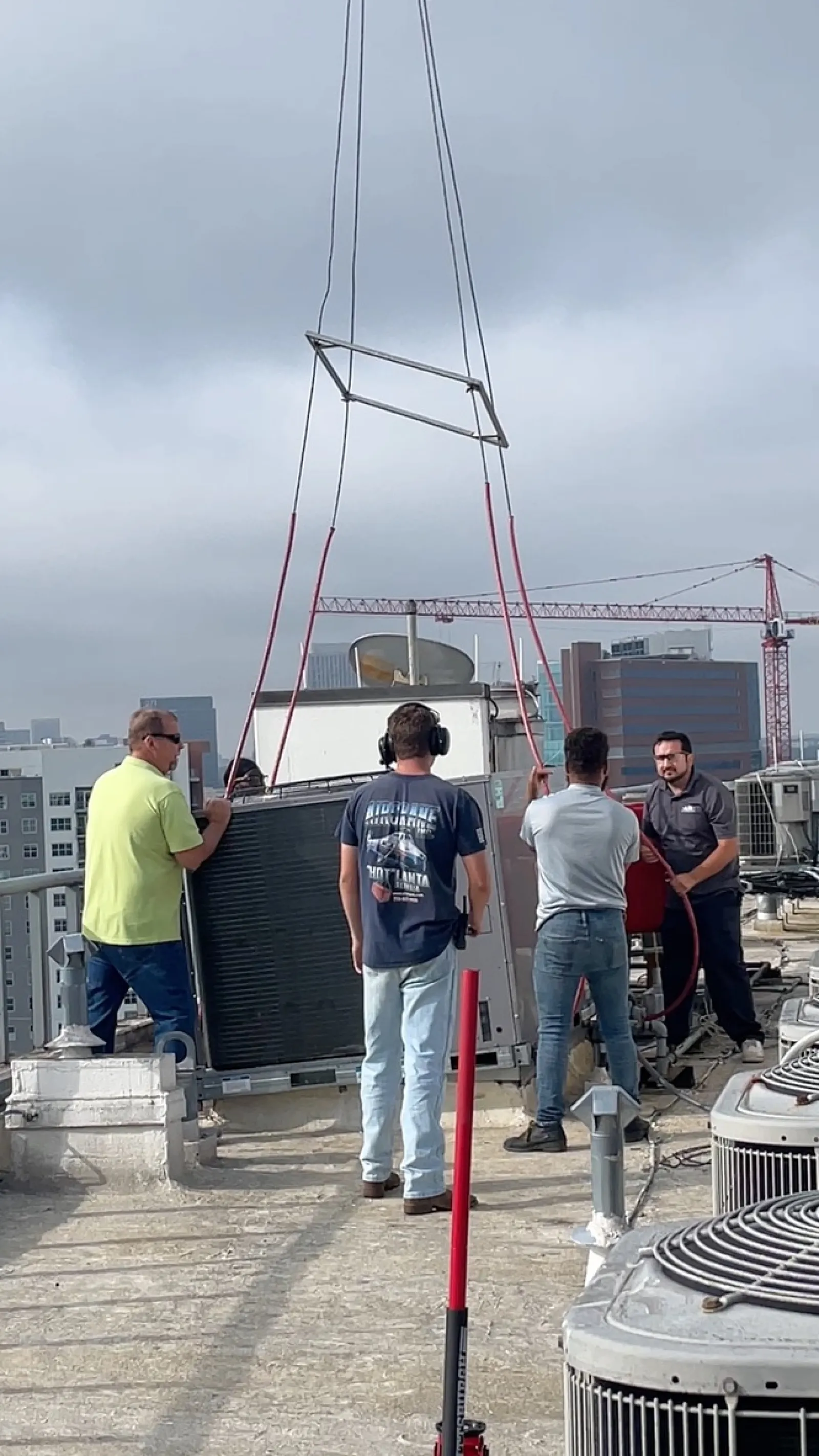 Four HVAC technicians guide a large unit on a rooftop using a crane under a cloudy sky in an urban area.