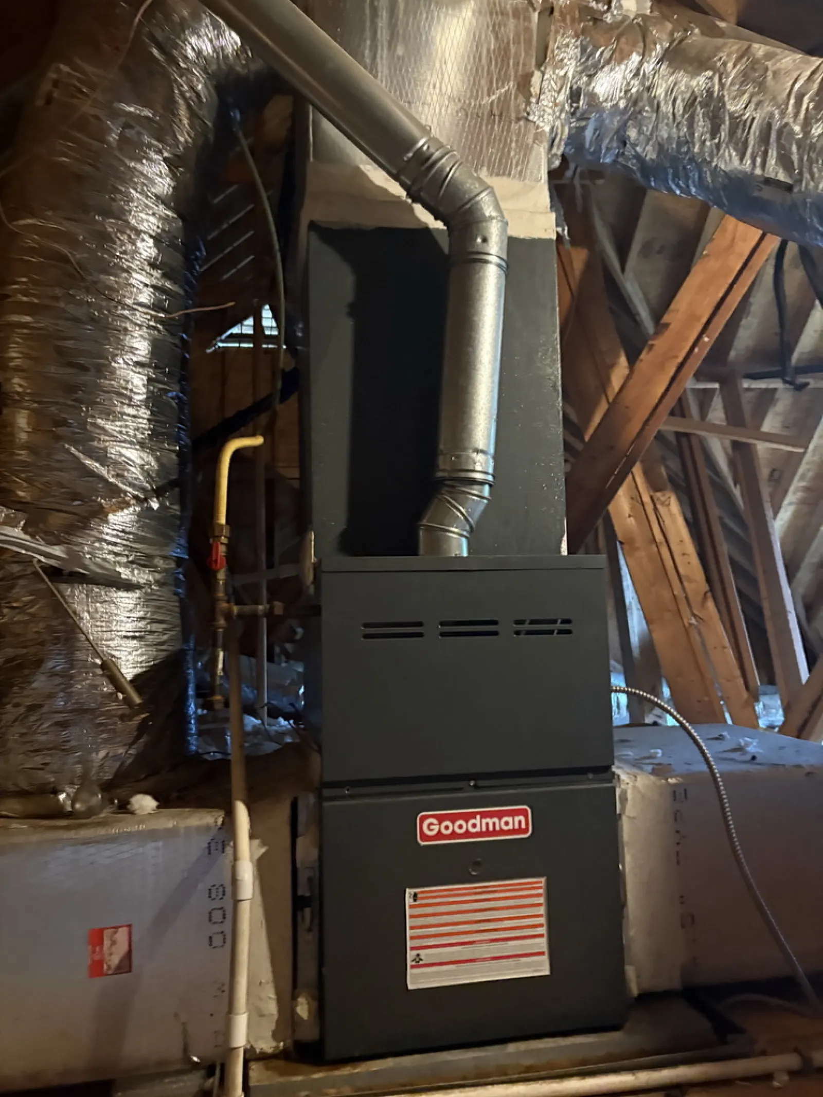 Goodman furnace installed in attic with insulated ductwork and exposed wooden beams around.