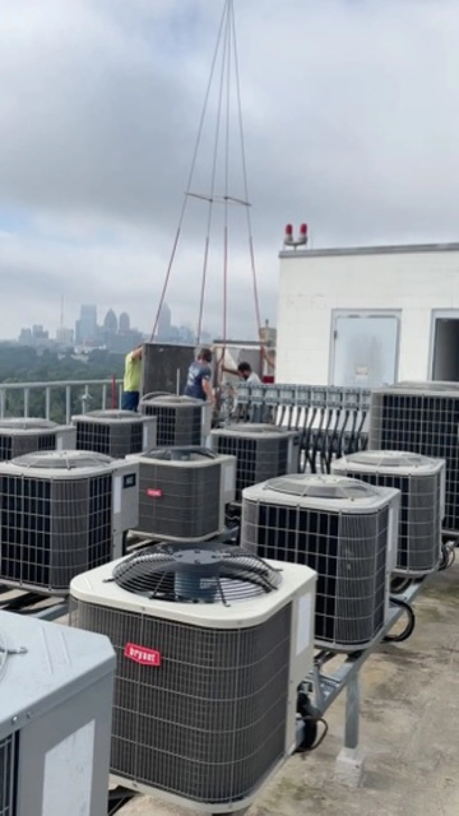 Technicians installing HVAC units on a rooftop with city skyline and cloudy sky in the background.