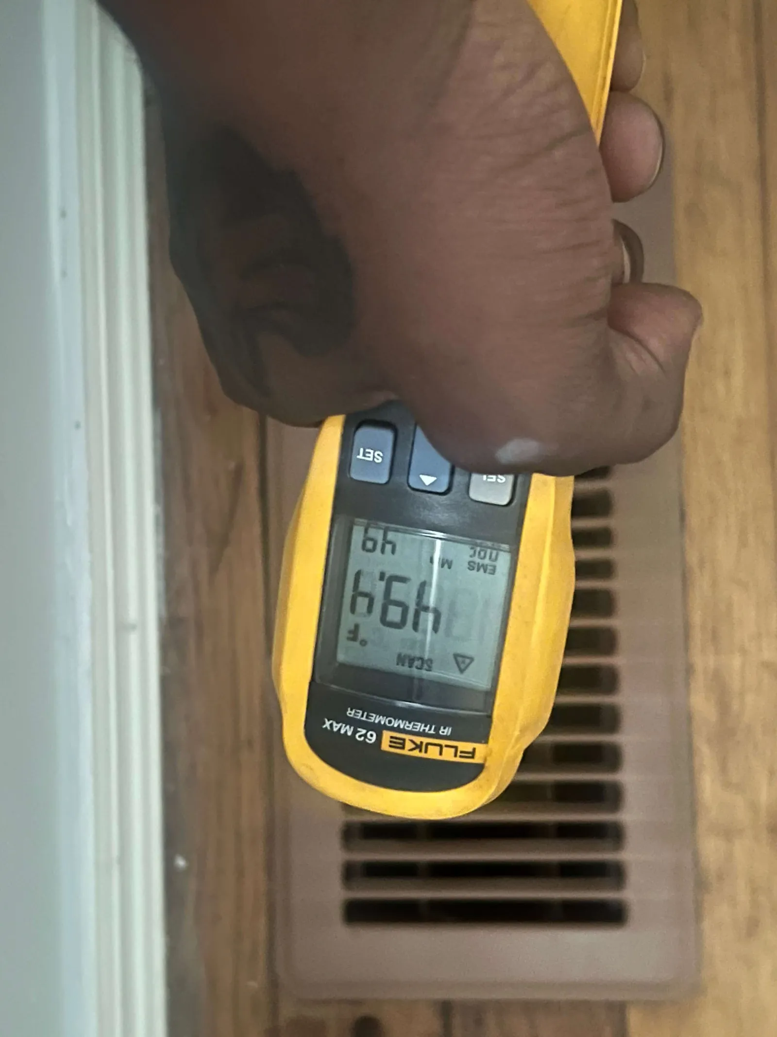 Hand holding a yellow Fluke 62 MAX IR thermometer measuring temperature near a floor vent showing 49.1°F.