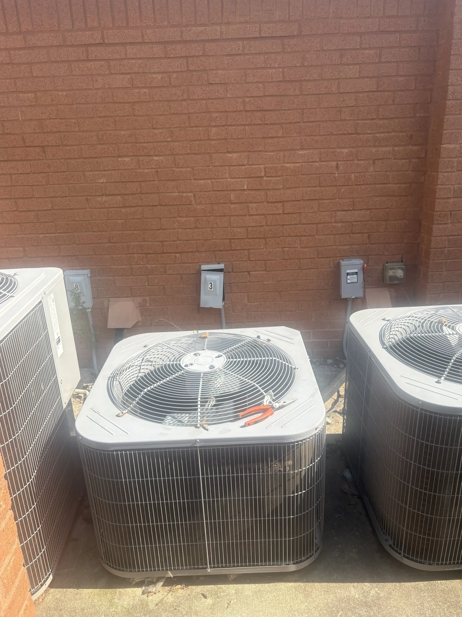 Three outdoor air conditioning units against a red brick wall with a pair of red pliers on the center unit's top grille.