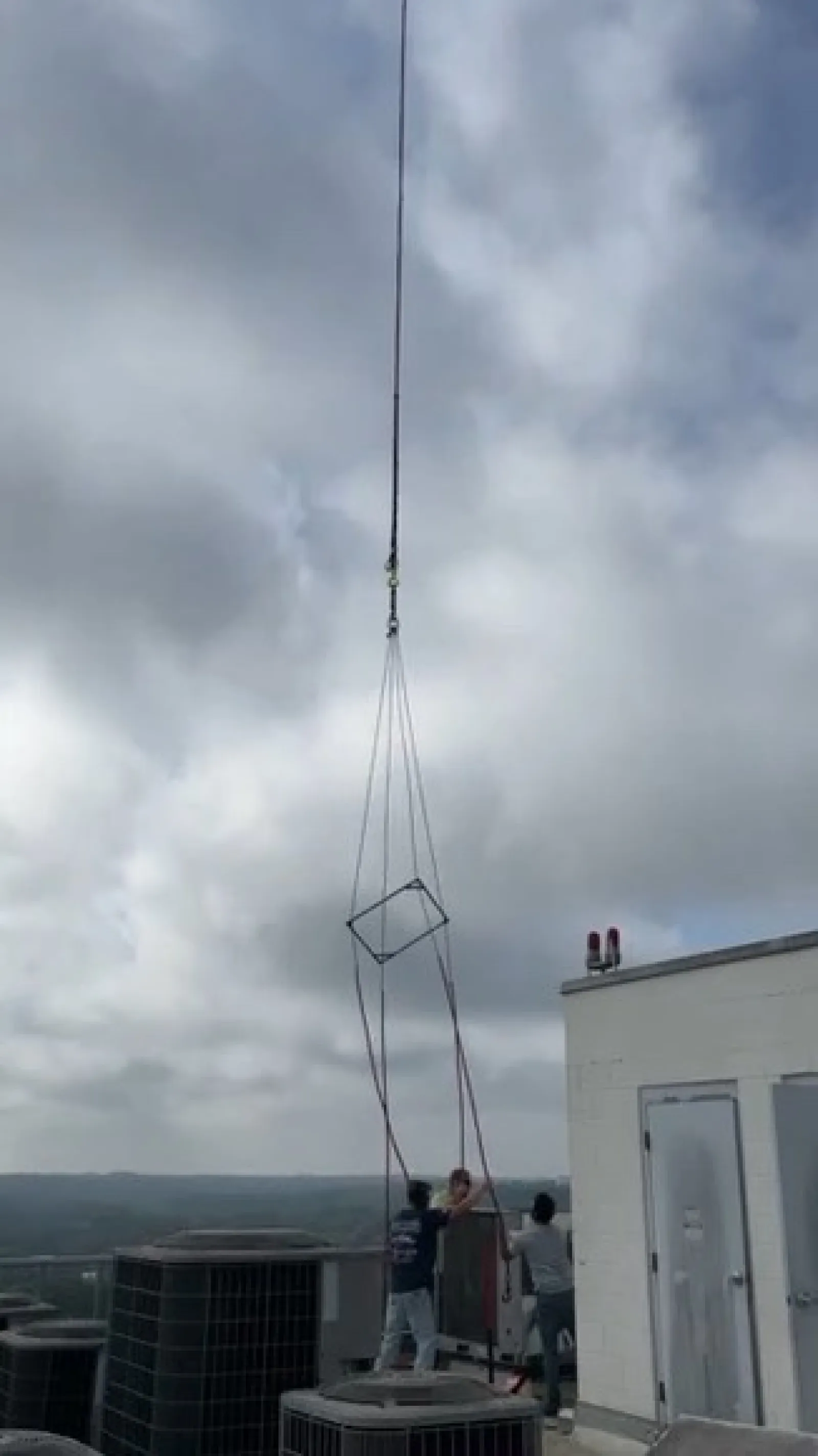 Two workers on rooftop guiding a large metal frame suspended by crane cables in cloudy weather.