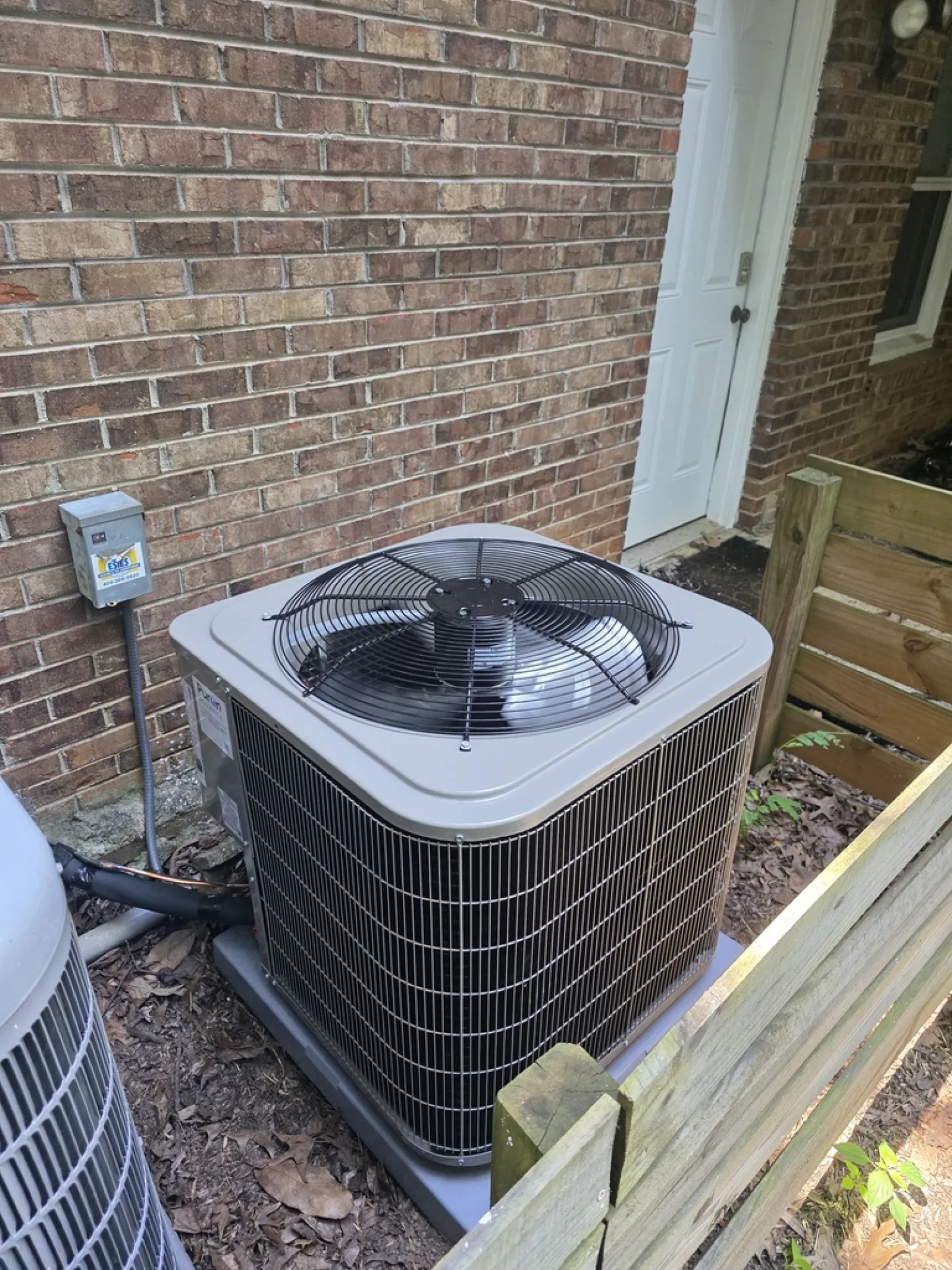 Outdoor air conditioning unit installed beside a brick house wall, surrounded by a wooden fence.