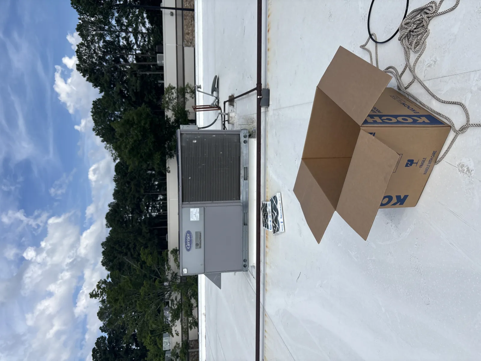 Open cardboard box and rooftop HVAC unit on a flat white roof under partly cloudy sky.
